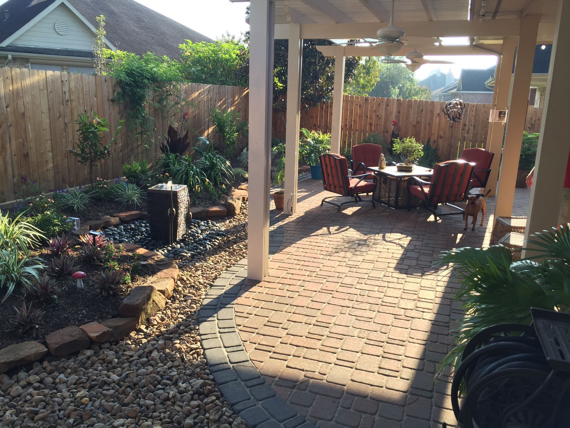 Brick patio walkway with red chairs under a covered pergola in a landscaped backyard garden