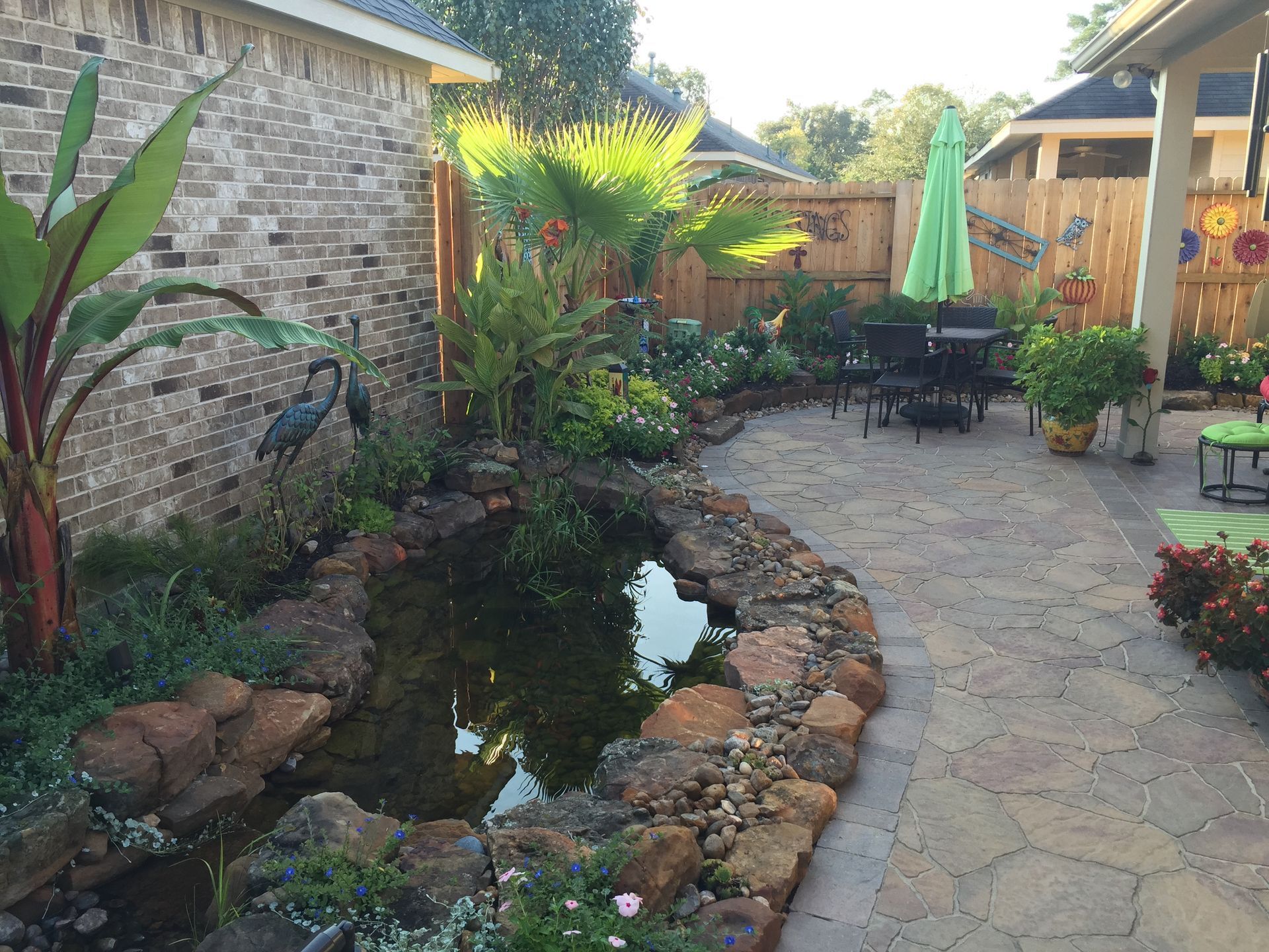 Backyard patio with a small pond, stone path, tropical plants, and outdoor seating under umbrellas