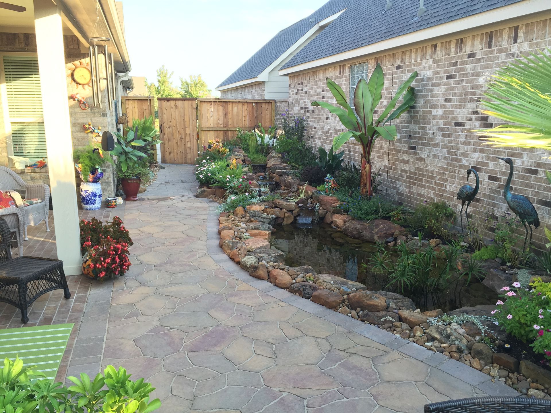 Stone garden path beside a brick house, with tropical plants and a small pond area.