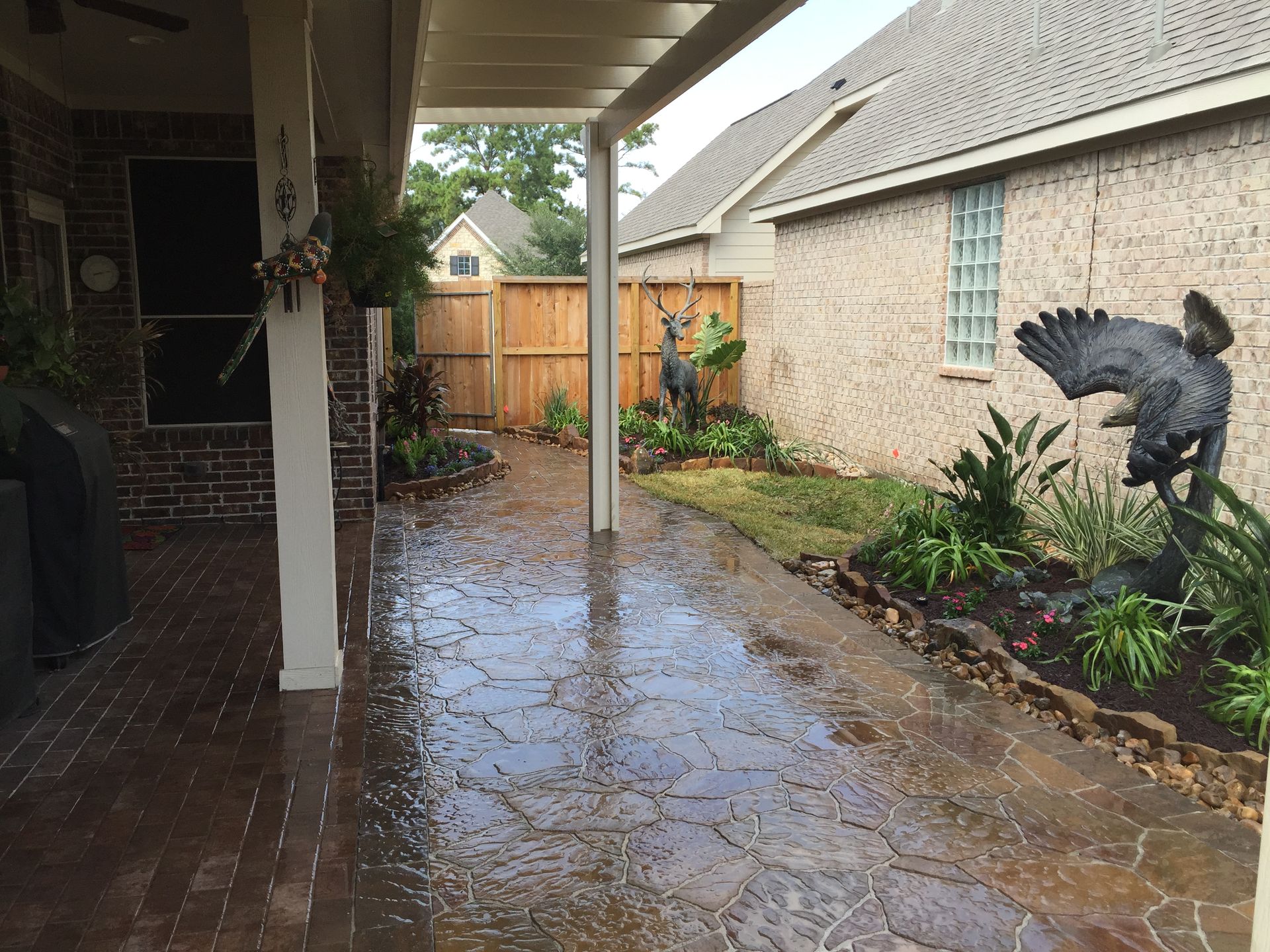 Rainy backyard patio with wet stone floor, covered porch, wooden fence, and garden statue.
