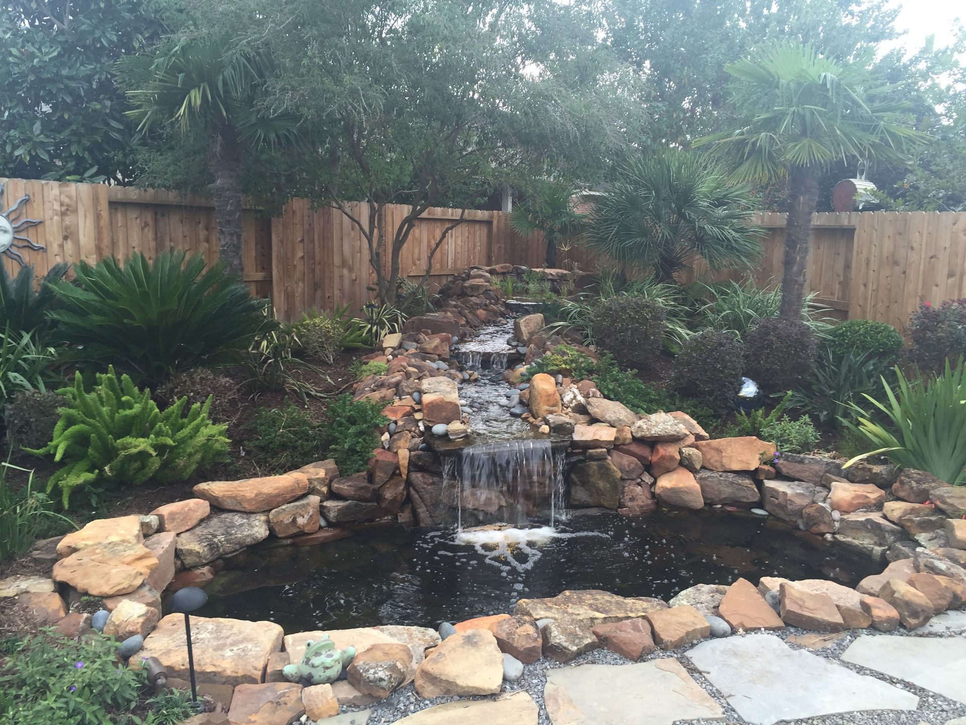 Backyard pond waterfall with rocks, greenery, and a wooden fence backdrop