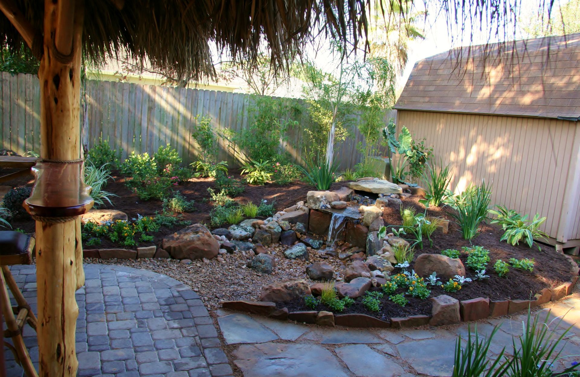 Backyard garden with stone path, rock-lined planter, and a small fountain beside a fence and shed.