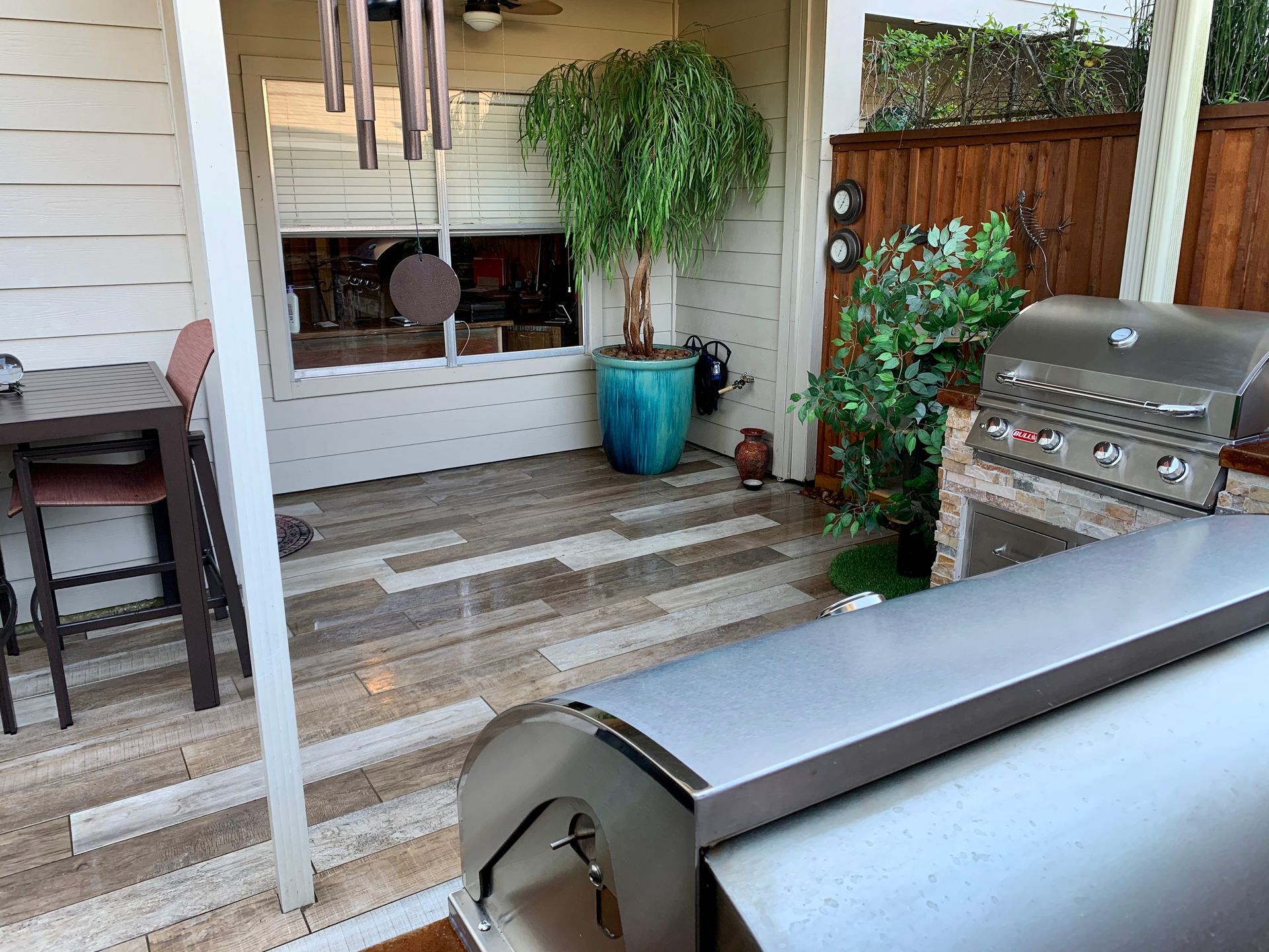 Covered patio with tiled floor, potted plant, small table, and stainless steel grill near wooden privacy fence.