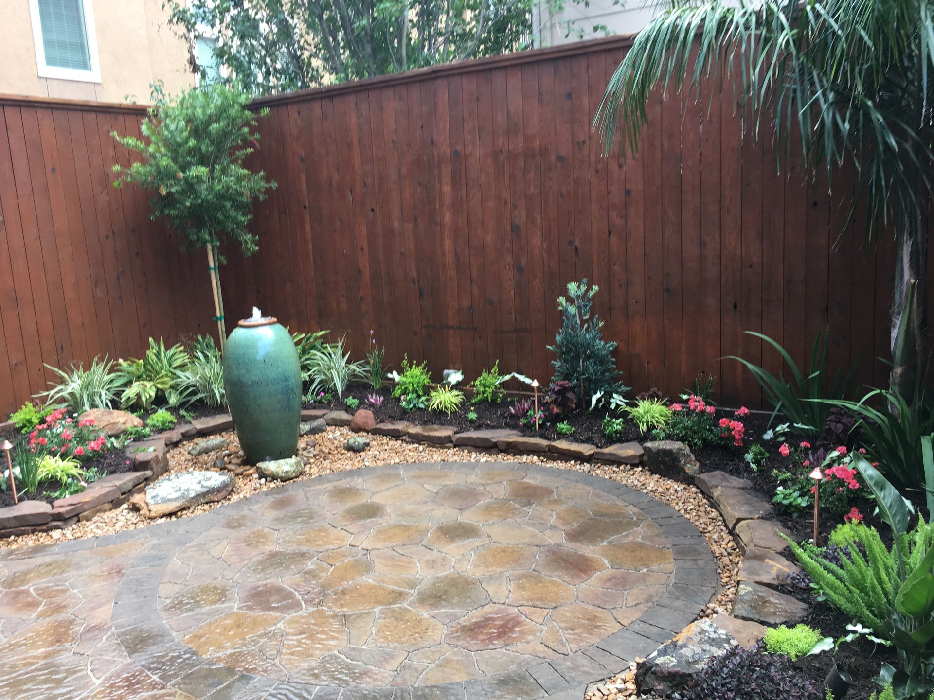 Small landscaped backyard patio with circular stone paving, potted plants, and a wooden fence