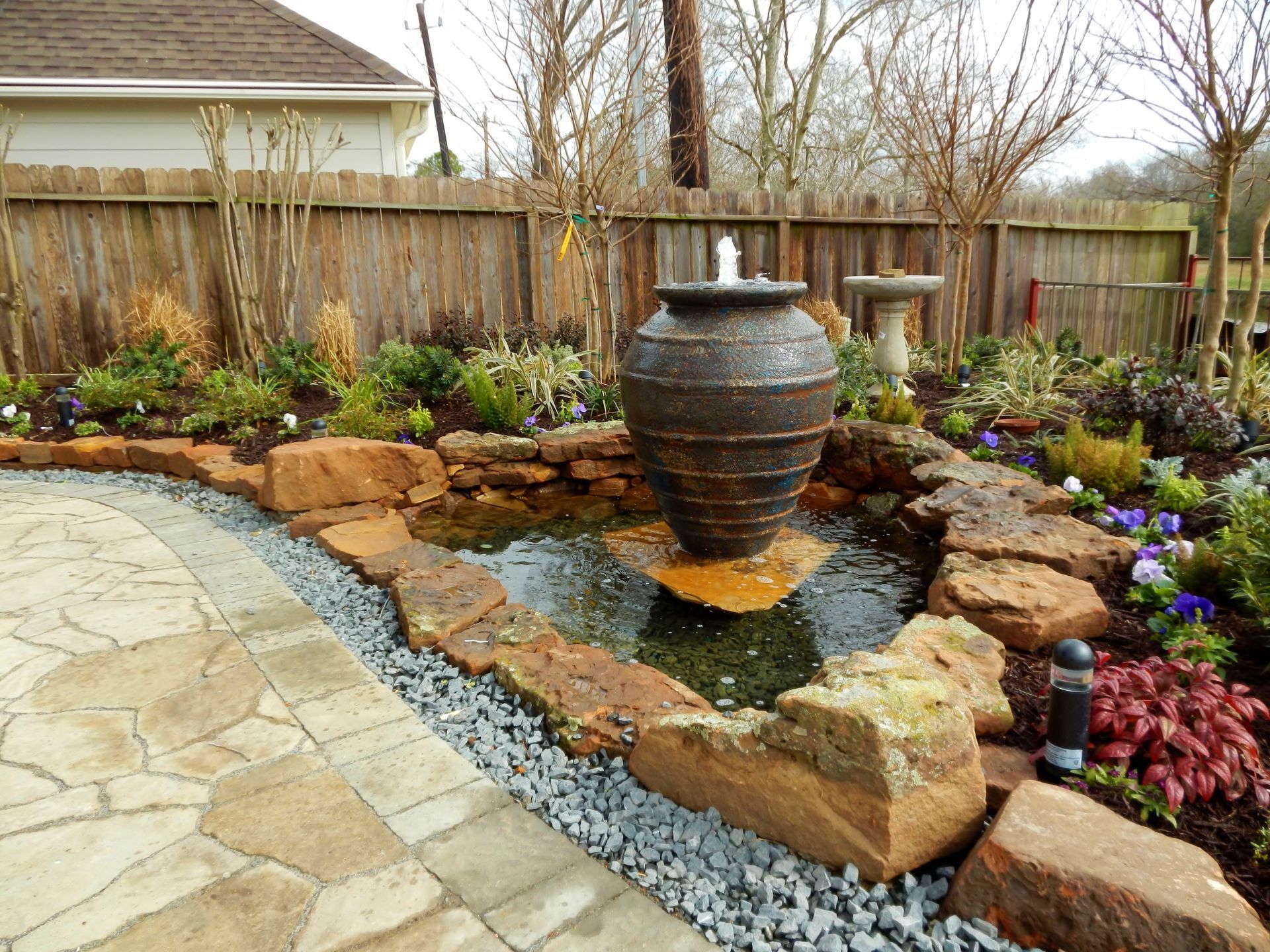 Backyard garden with a large ceramic water urn fountain, stone edging, and blooming flowers