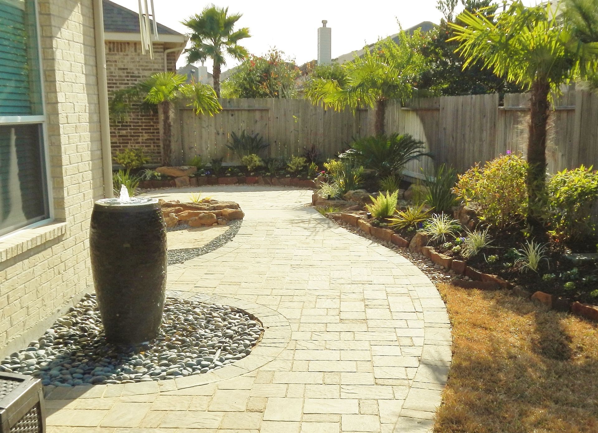 Curved stone patio walkway beside a house, with a large planter and landscaped backyard fence line