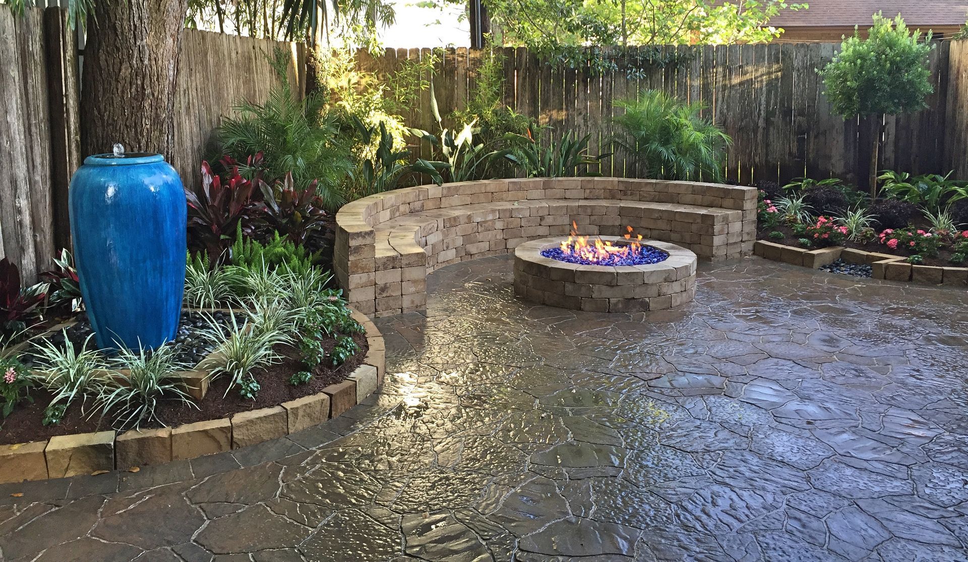 Rain-soaked stone patio with a blue urn planter and a circular fire pit in a landscaped garden