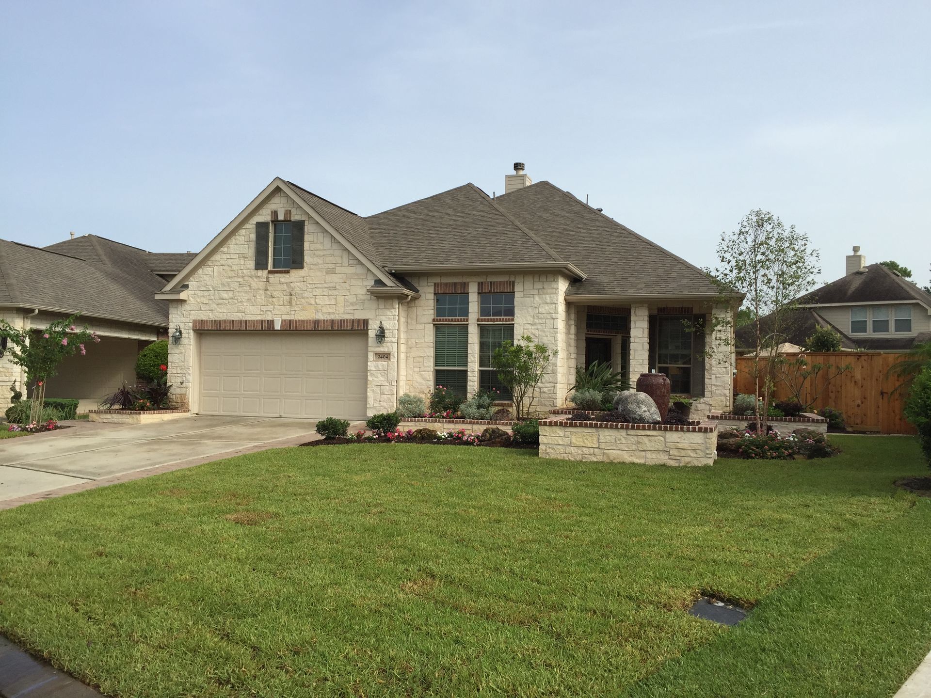 Suburban brick house with a two-car garage, green lawn, and a landscaped front yard under a clear sky