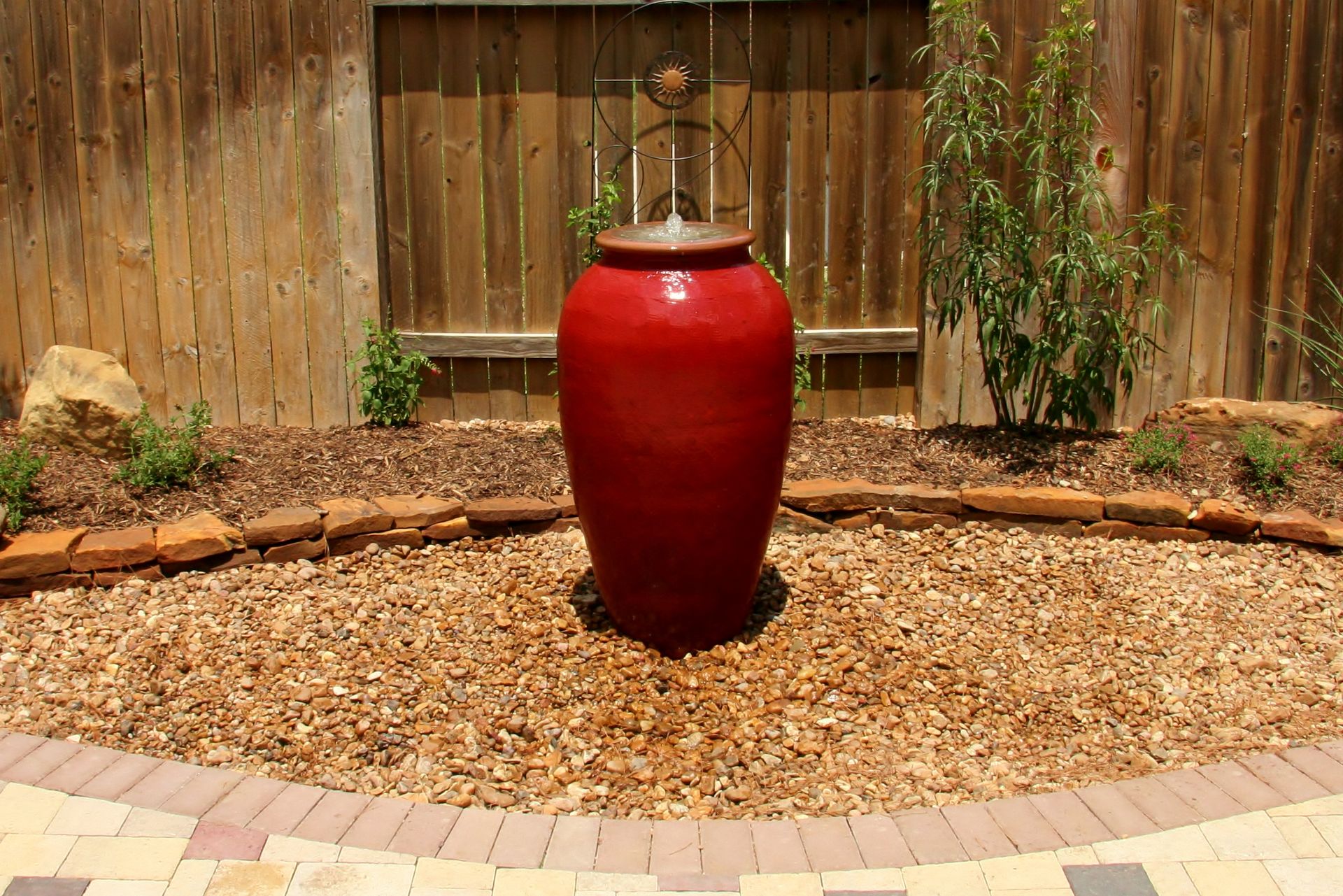 Red ceramic garden urn centered in a gravel bed against a wooden fence.