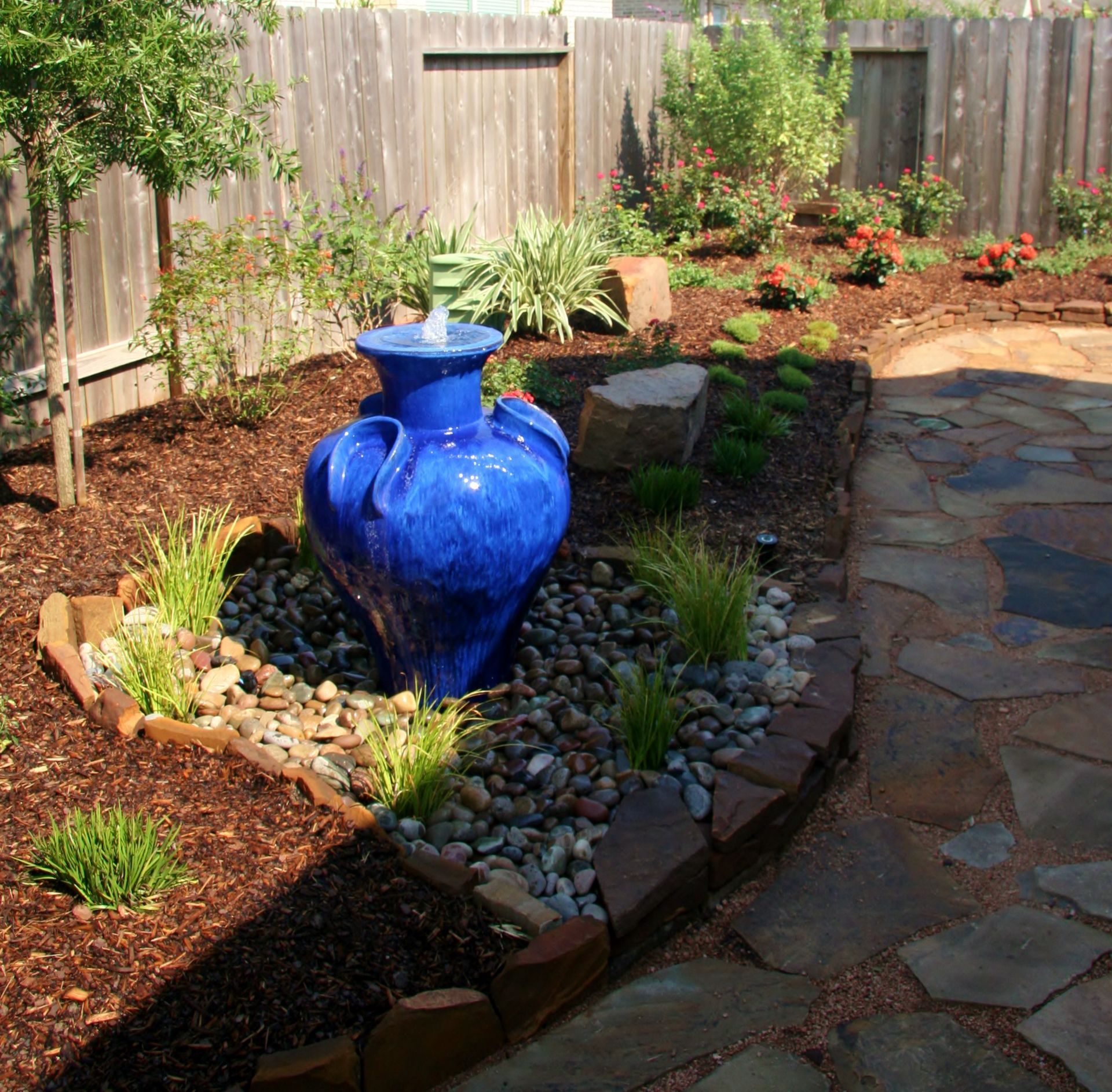 Backyard garden with a blue ceramic fountain, stone path, and mulched flower beds