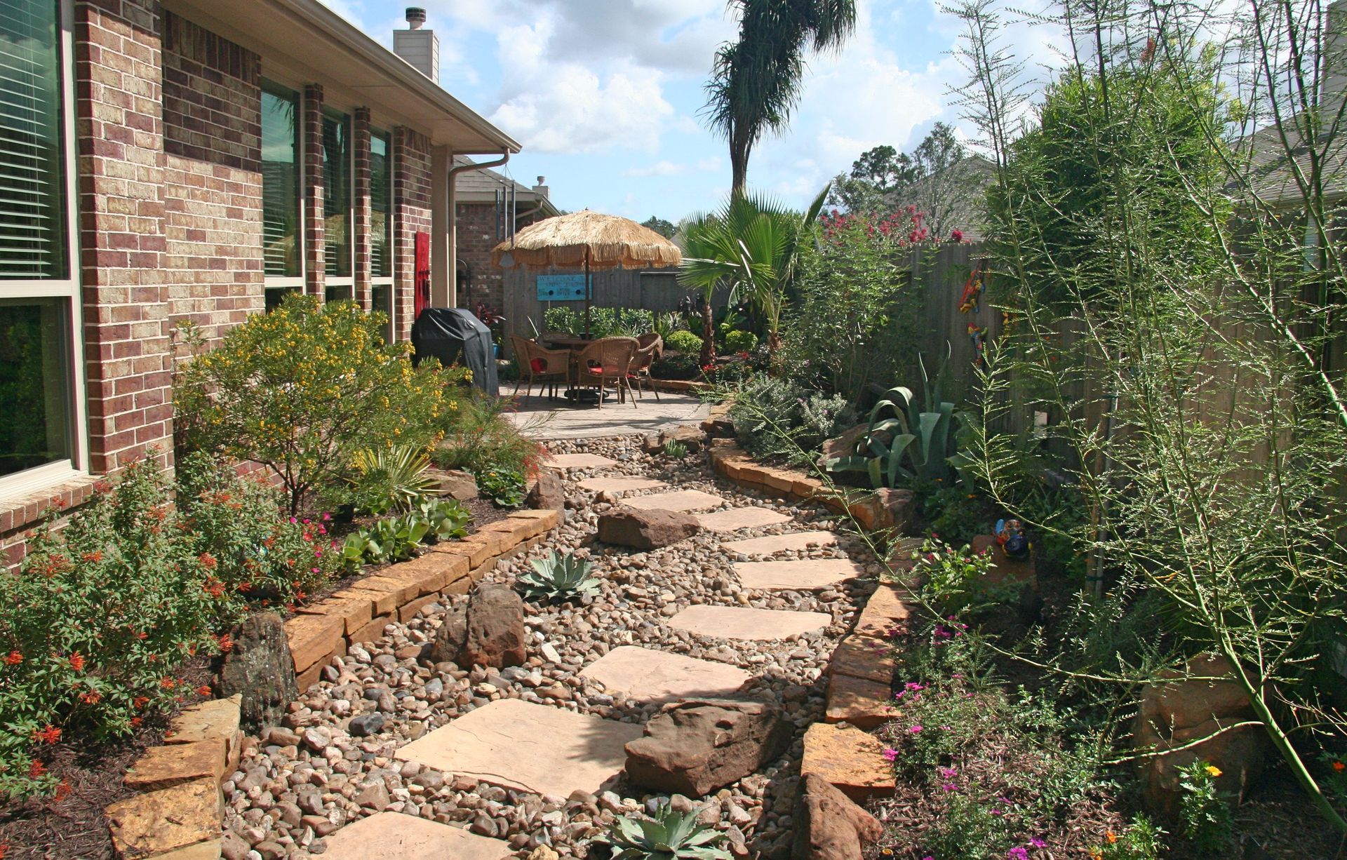 Stone garden path beside a brick house, bordered by flowers and greenery on a sunny day