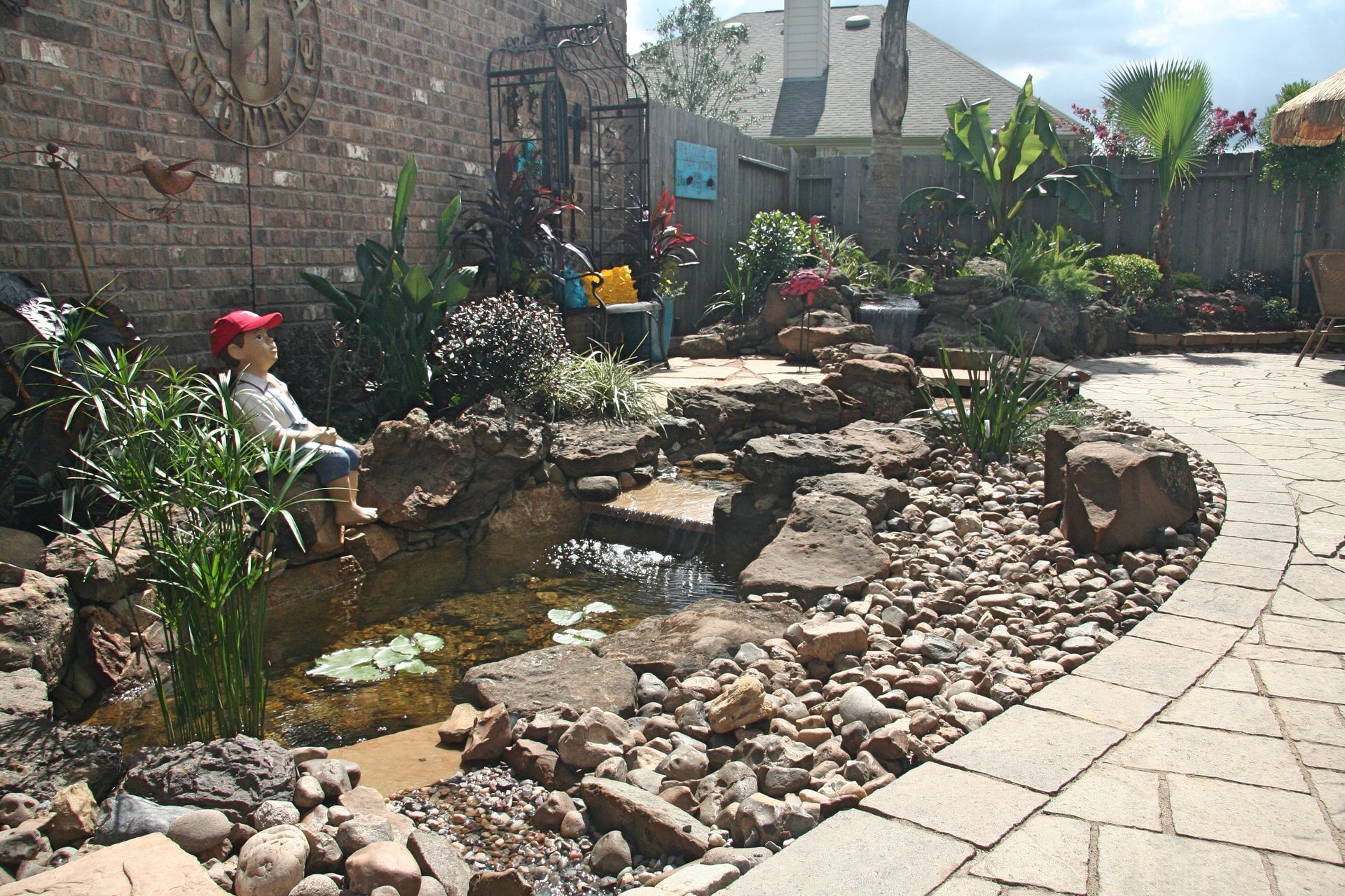 Backyard pond with rocks, a small waterfall, and a curved stone patio in bright sunlight
