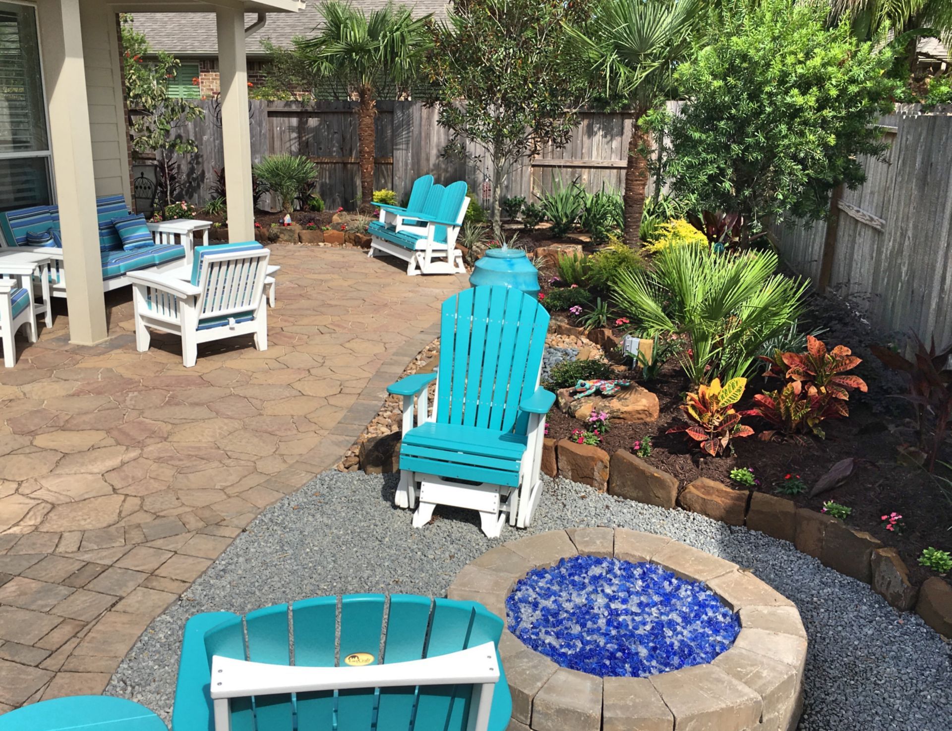Patio with turquoise Adirondack chairs, stone paving, and a blue-tiled fire pit beside lush garden plants