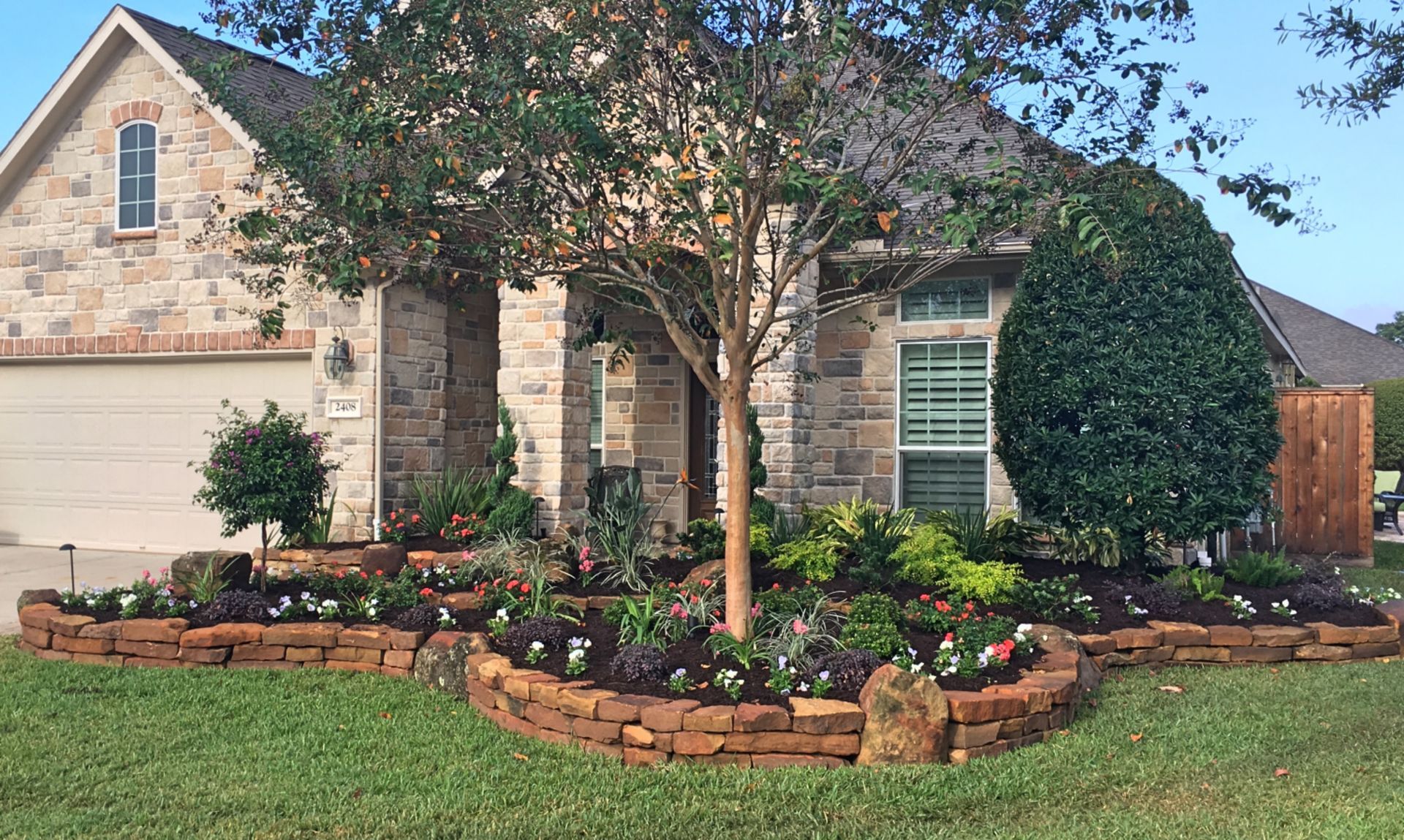 Stone house with a manicured front yard, brick flower beds, and large trees in the landscaping.