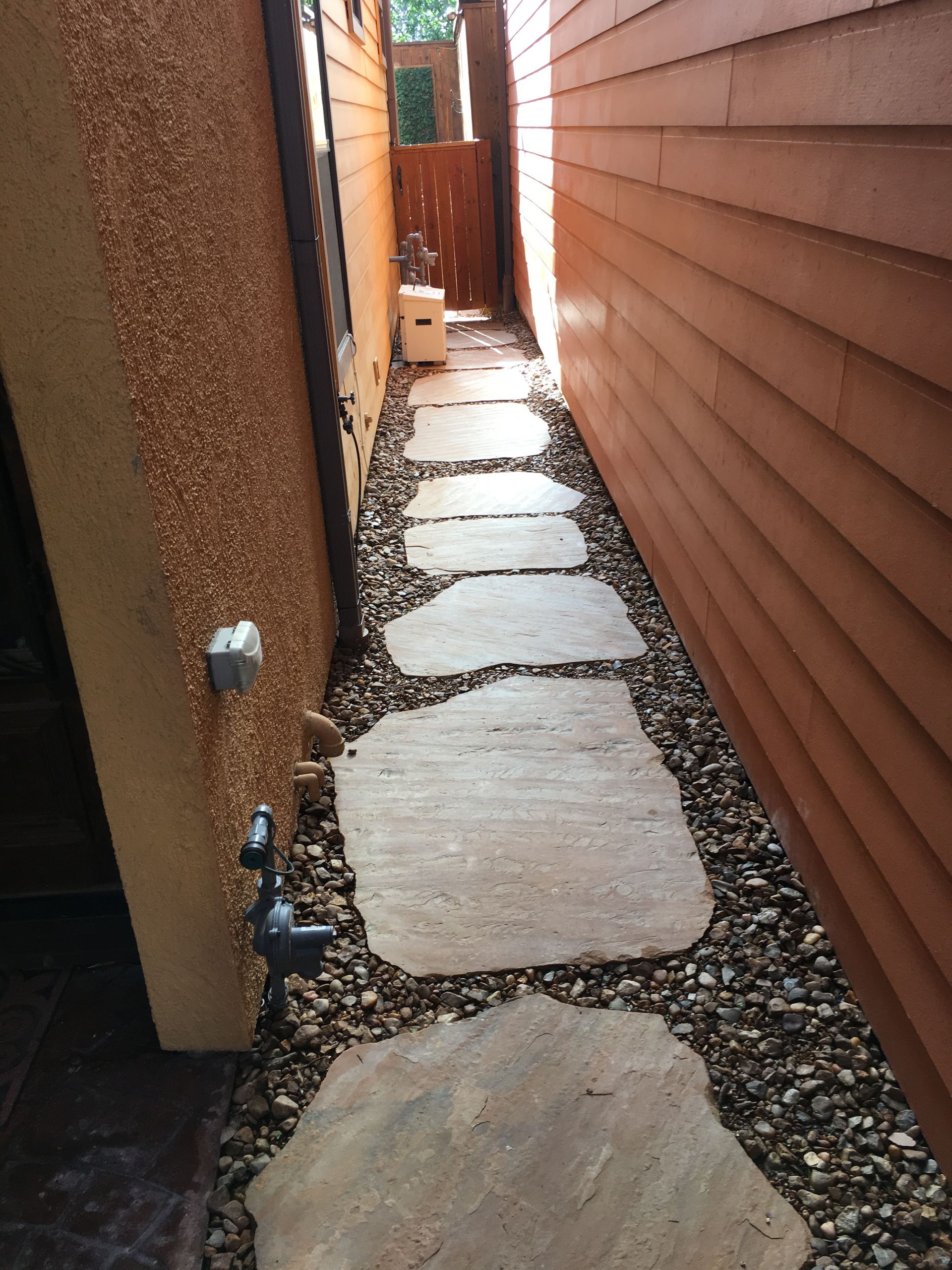 Narrow outdoor side passage with stepping stones between a stucco wall and brown siding.