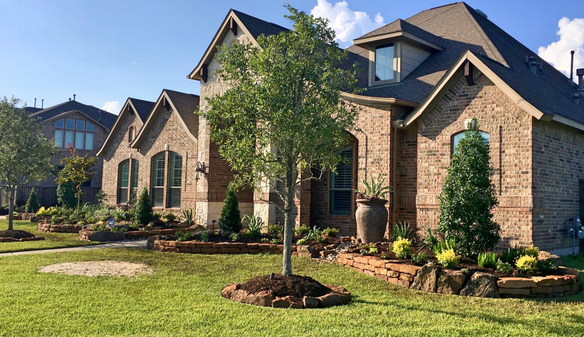 Large brick suburban house with gabled roof, front porch, and landscaped yard with trees and shrubs.