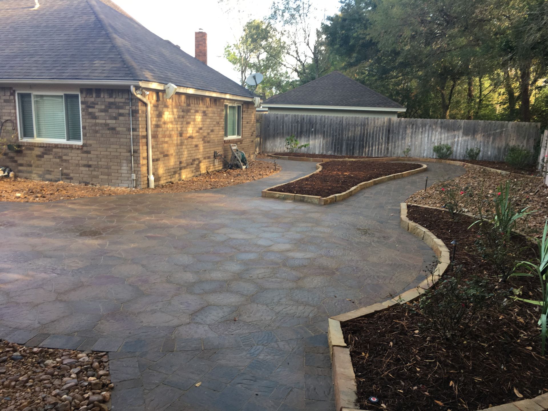 Curved concrete driveway beside a stone house, with fresh mulch beds and a white fence.
