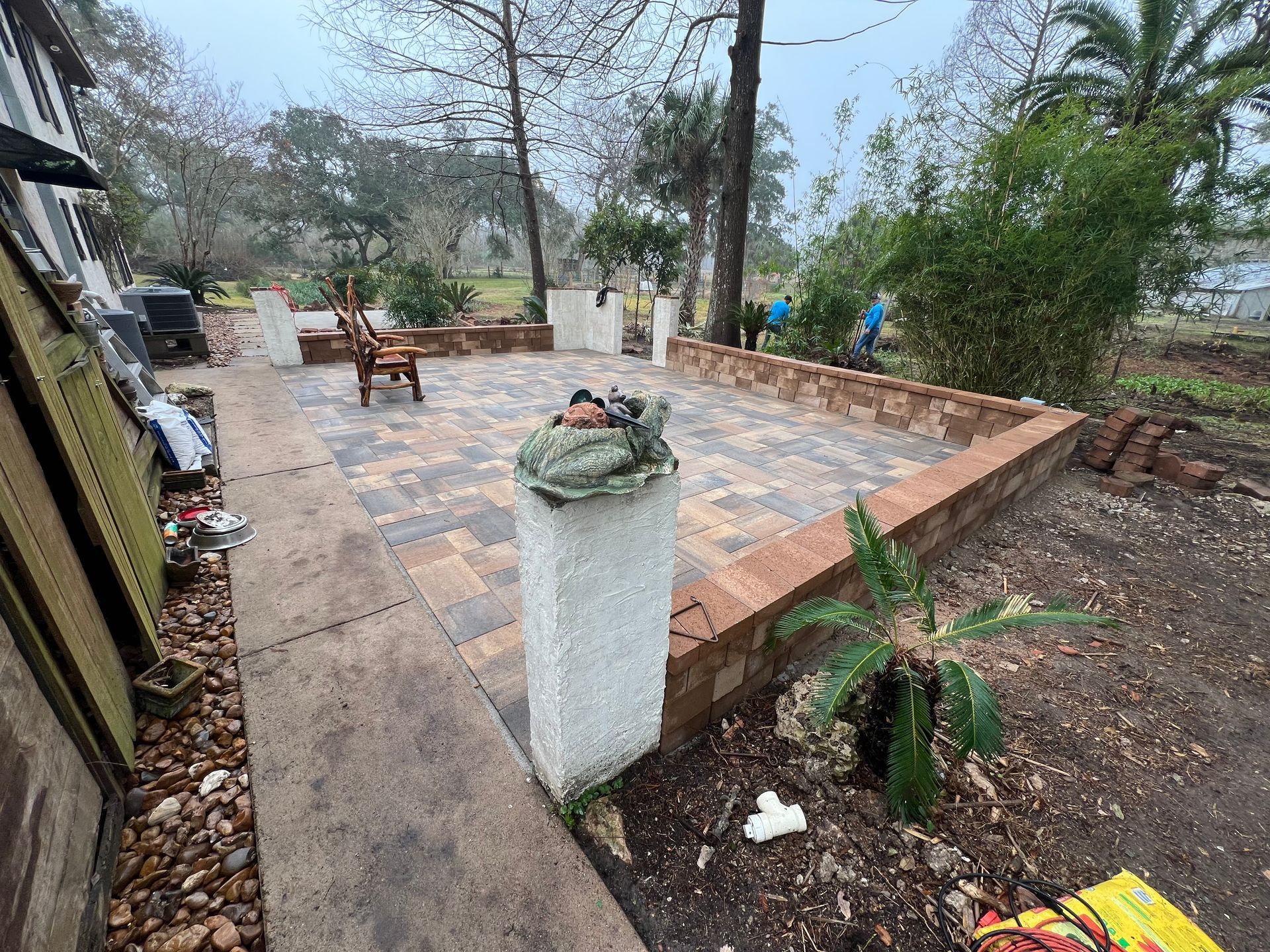 Outdoor patio with stone paving, benches, potted plant, and garden beds beside trees and a road.