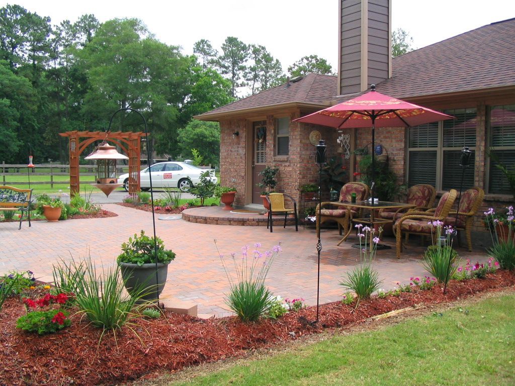 Outdoor patio with tables, umbrellas, potted flowers, and a brick building beside a parking area.