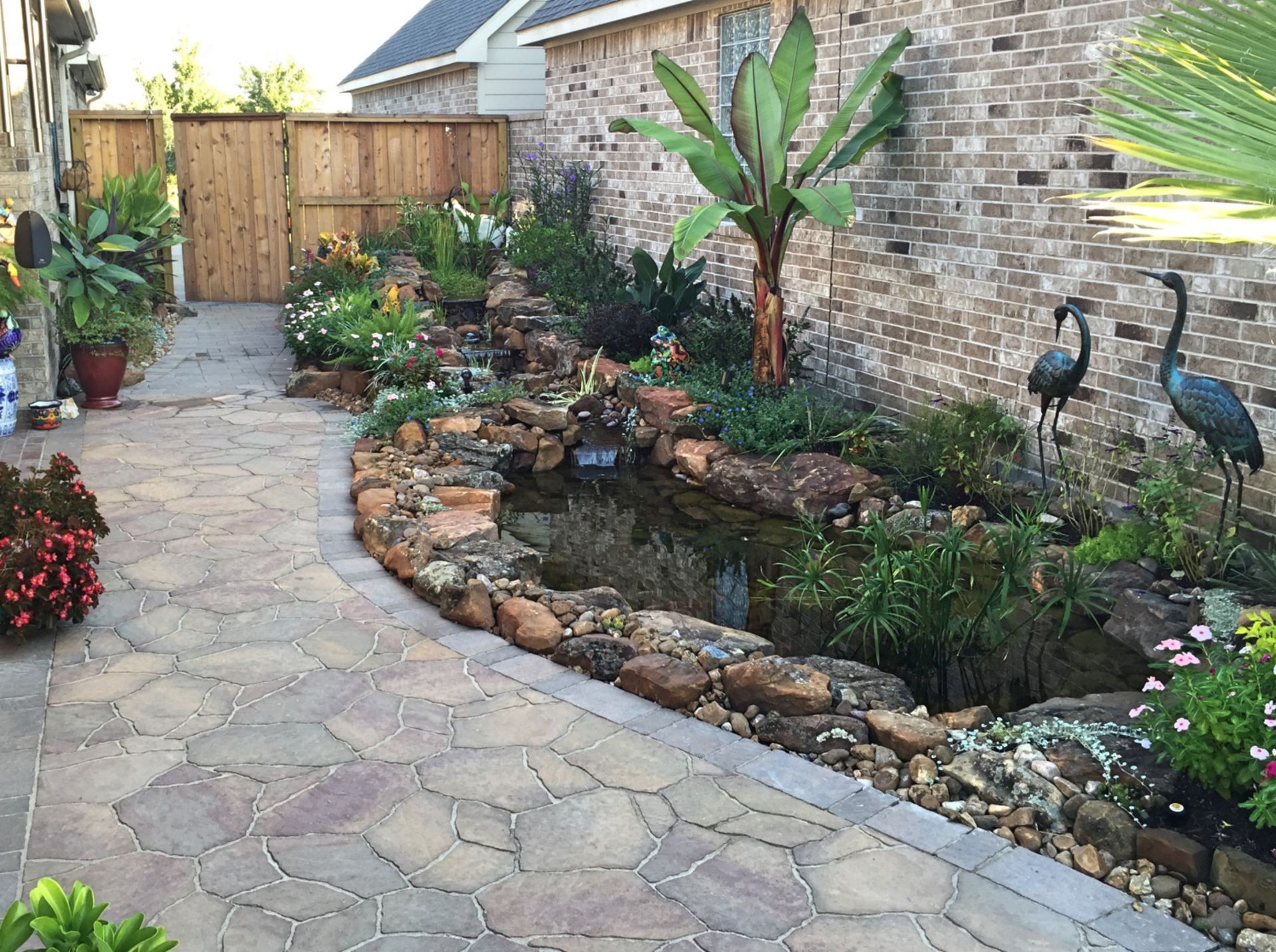 Backyard garden with stone path, pond, potted plants, and two decorative bird sculptures by a brick wall