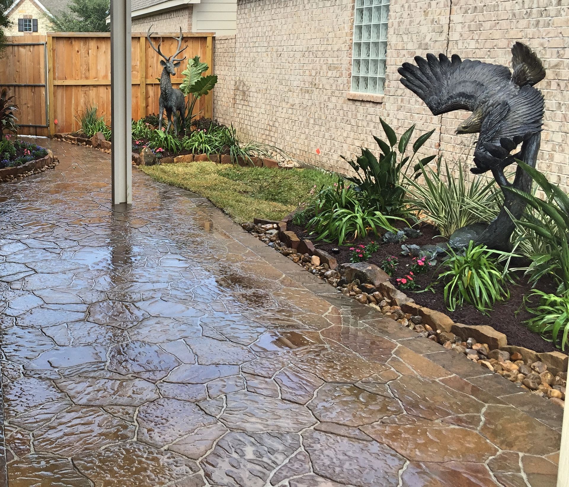 Rain-soaked stone patio beside a garden, with a black bird sculpture and wet reflections.