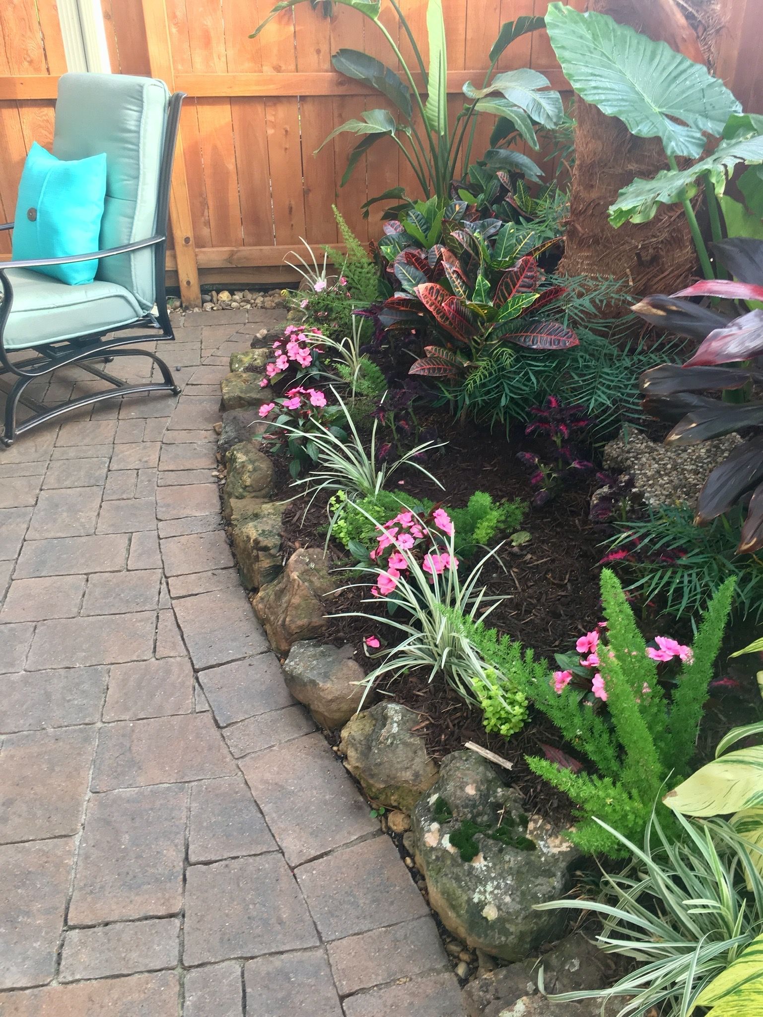 Brick patio beside a wooden fence, with a lush flower bed and turquoise chairs in the corner.