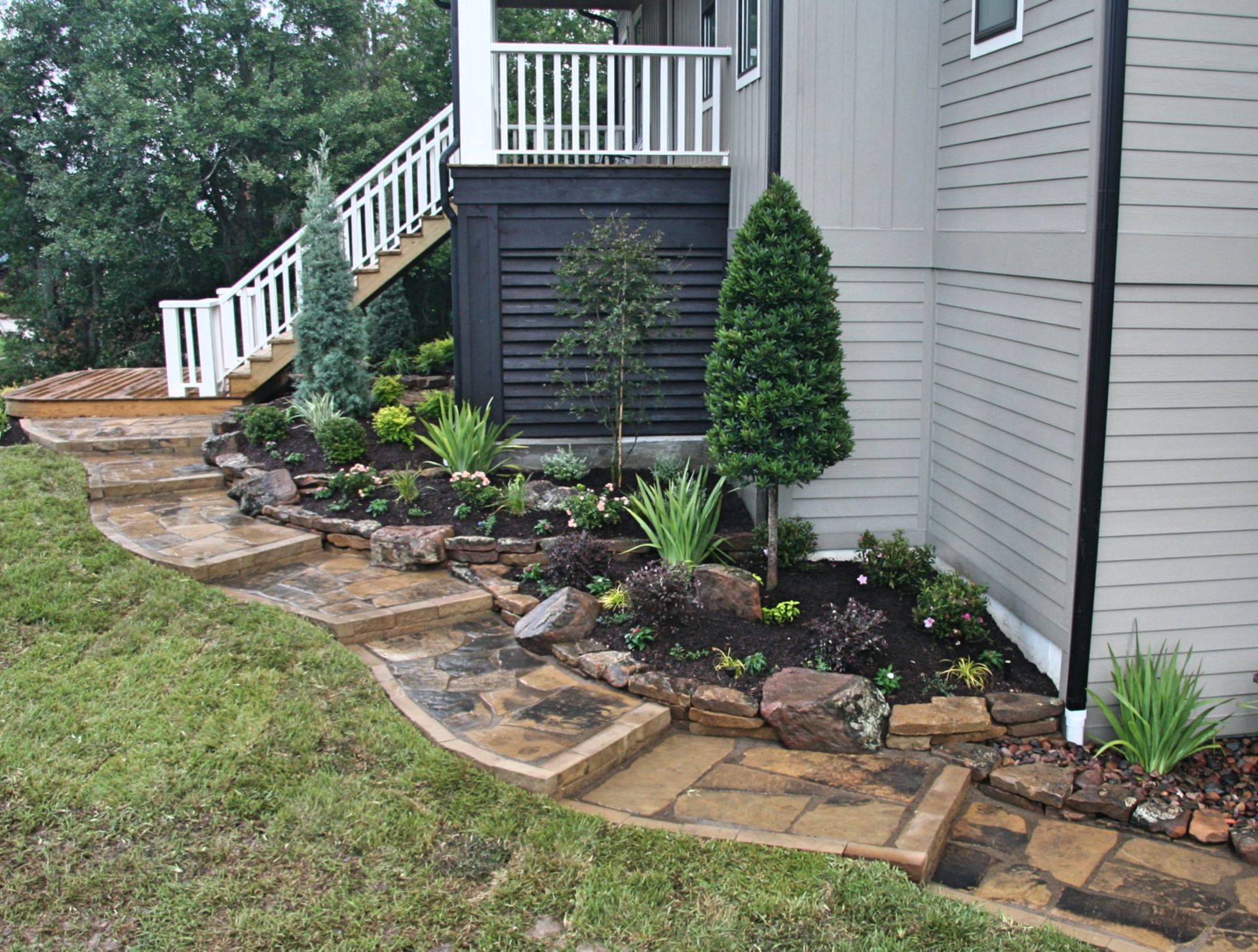 Curved stone flower bed with shrubs and small trees along a house and porch