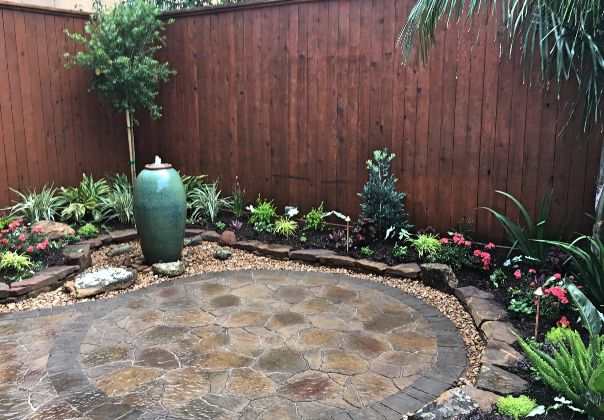 Stone patio courtyard with flower beds, wooden fence, and a tall green vase fountain
