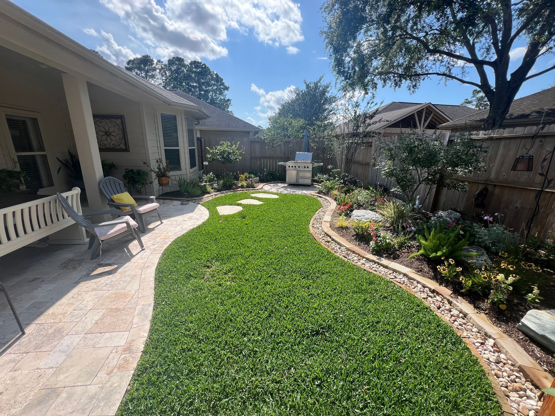 Curved green lawn beside a house with stone path, porch chairs, flower beds, and trees under a blue sky
