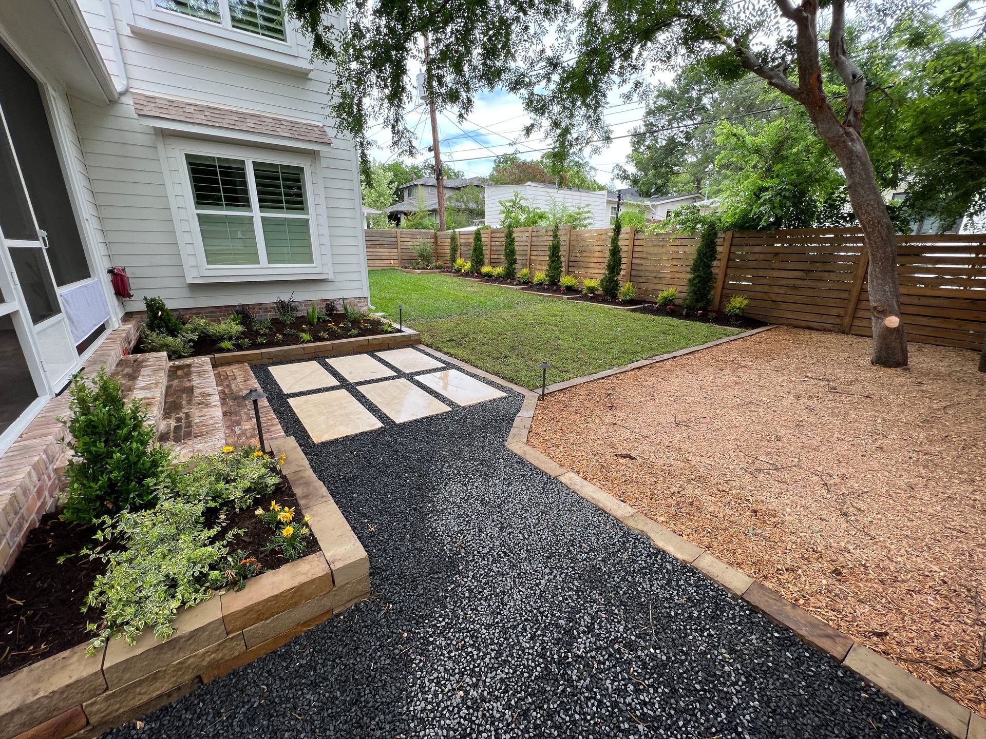 Landscape path with stepping stones, black gravel, raised garden bed, and tree in a backyard beside a house