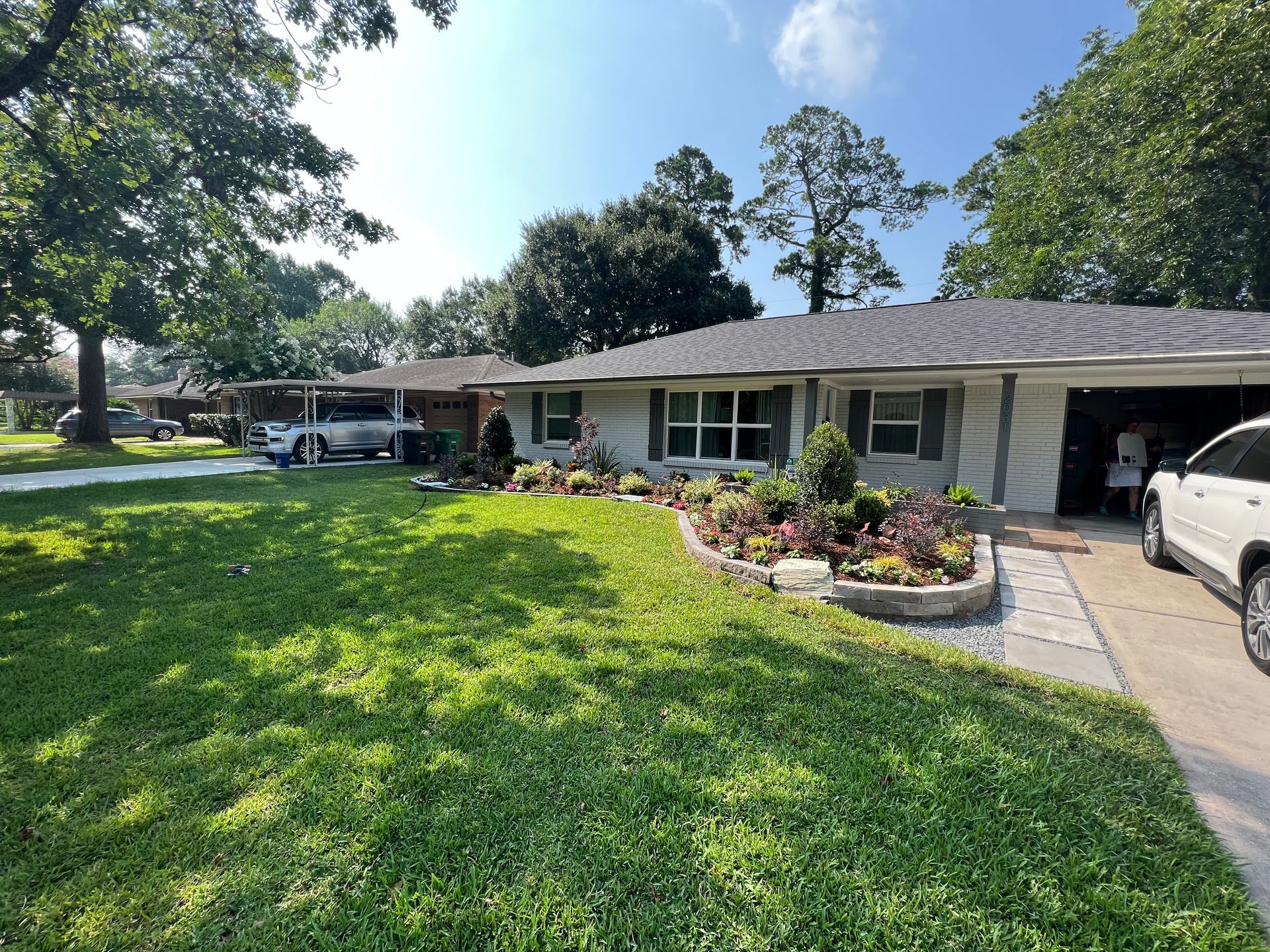 Single-story house with a green lawn, flower beds, and parked cars in a sunny suburban yard