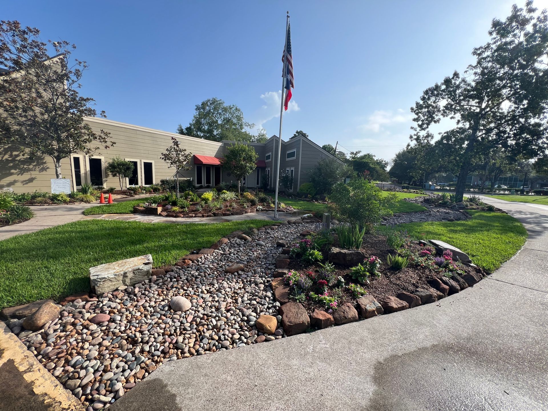 Landscaped building entrance with a flagpole, stone beds, and curved walkway under a blue sky