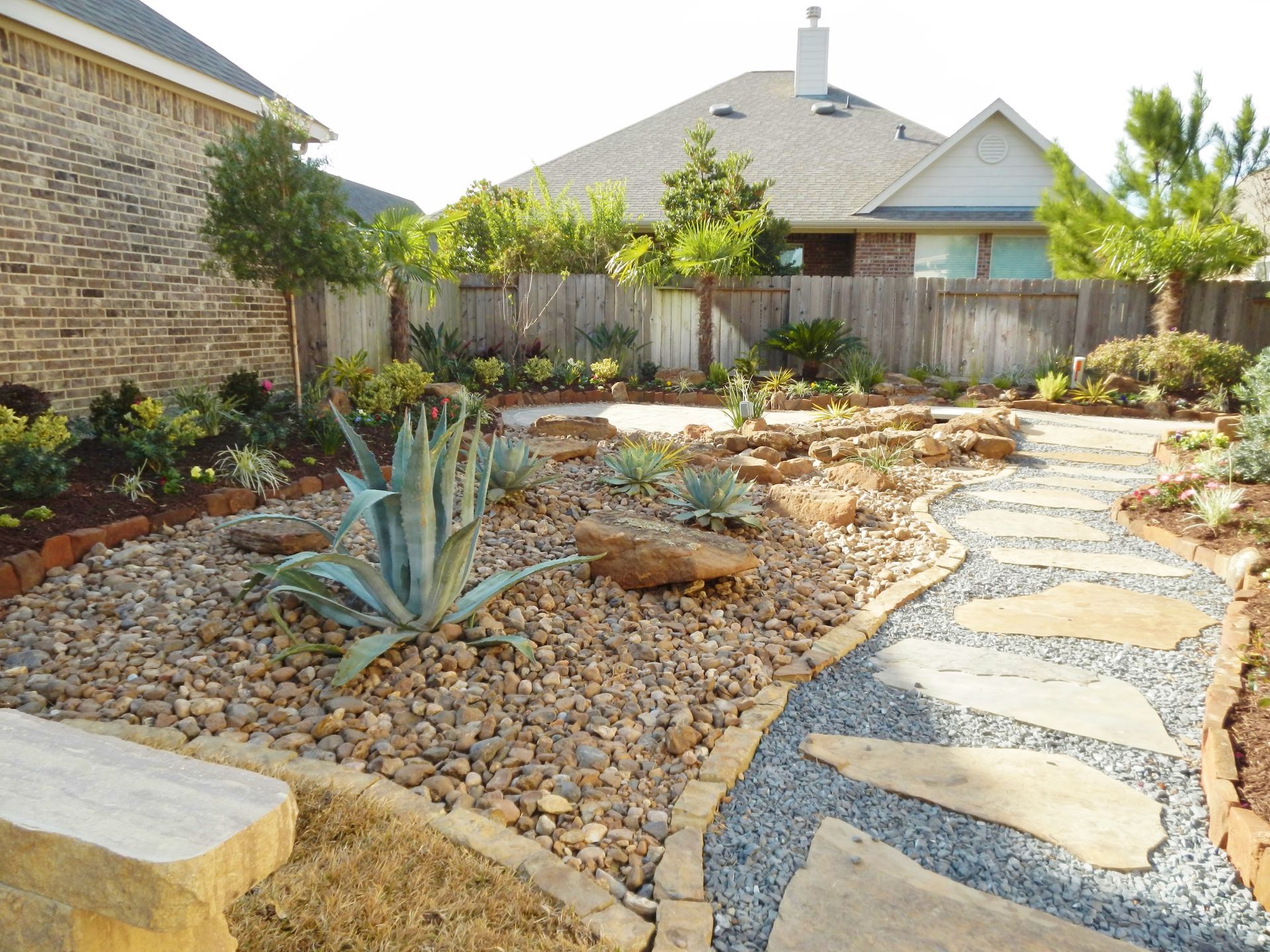 Desert-style backyard with stone path, agave plants, gravel beds, and wooden fence behind a house