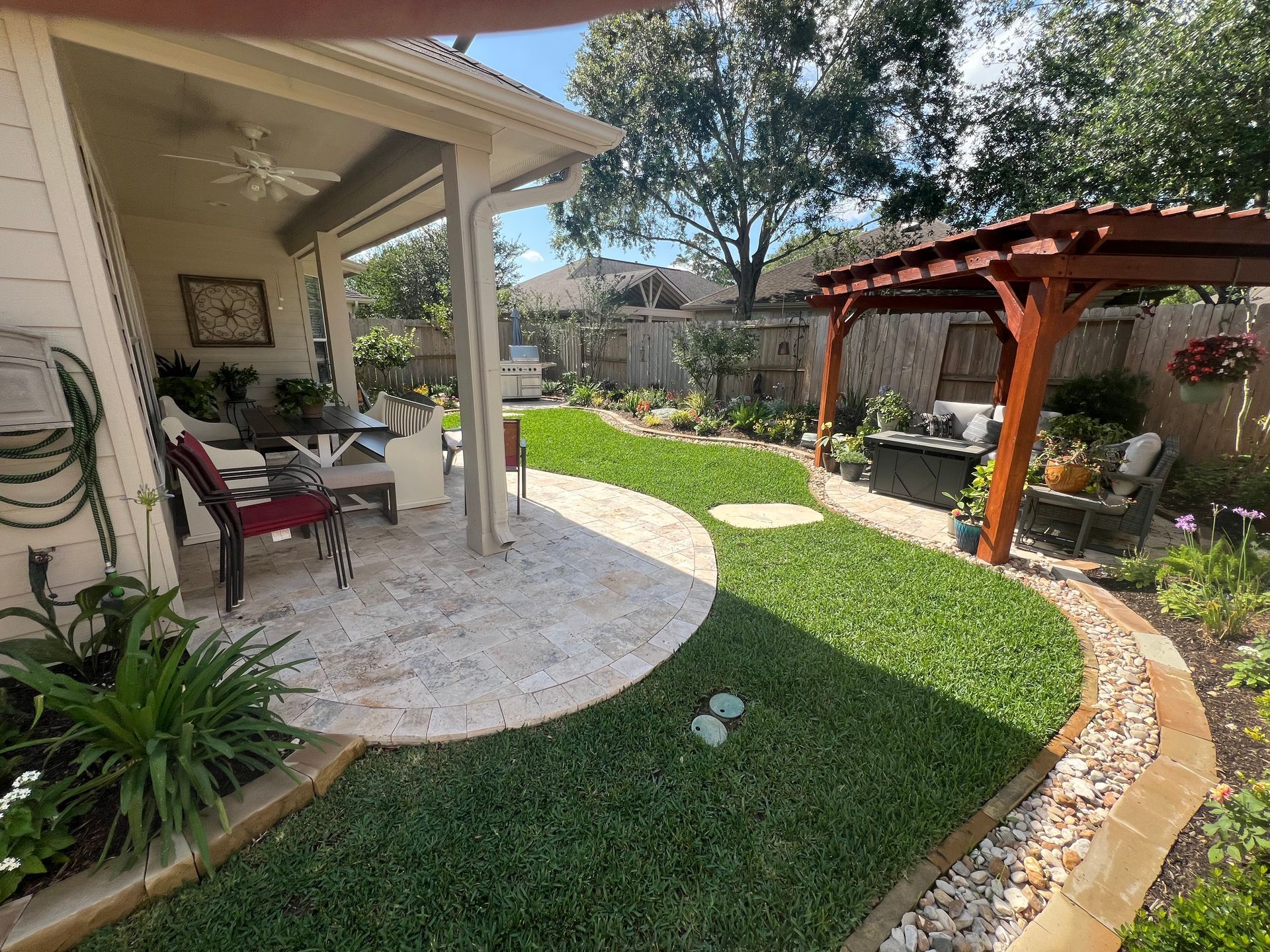 Backyard patio with covered seating, green lawn, curved stone path, and wooden pergola near a garden.