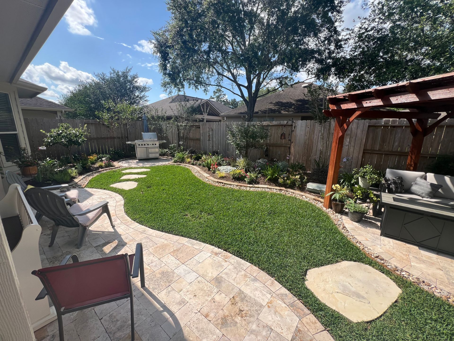 Backyard patio with curved stone path, green lawn, lounge chairs, pergola, and garden beds under a blue sky