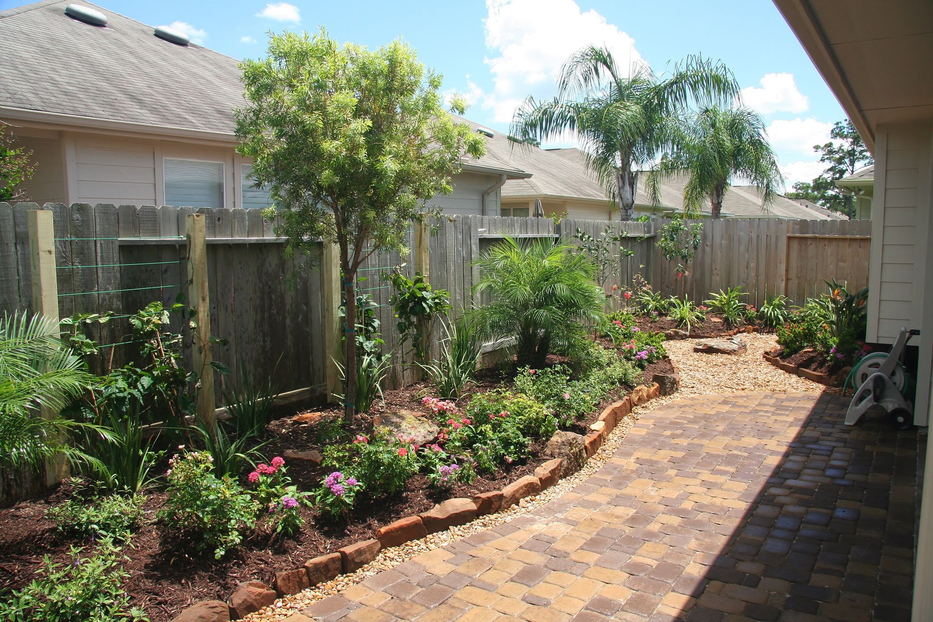 Brick-paved garden path beside a wooden fence and flowering plants in a sunny backyard