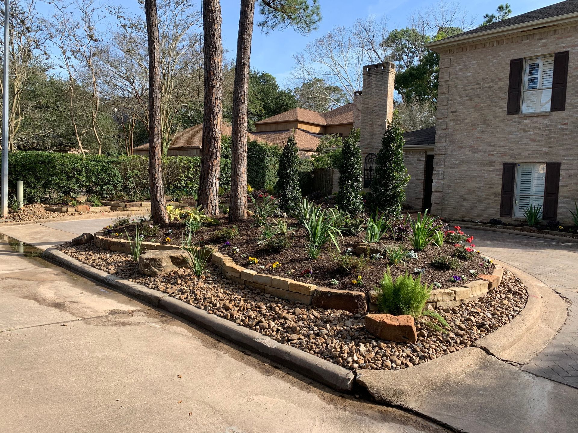 Landscaped courtyard with trees, flower beds, and a brick building beside a paved driveway