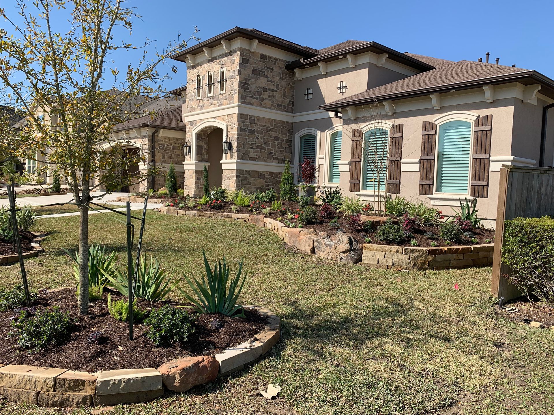 Mediterranean-style house with stone facade, teal shutters, and a landscaped front yard under a sunny sky