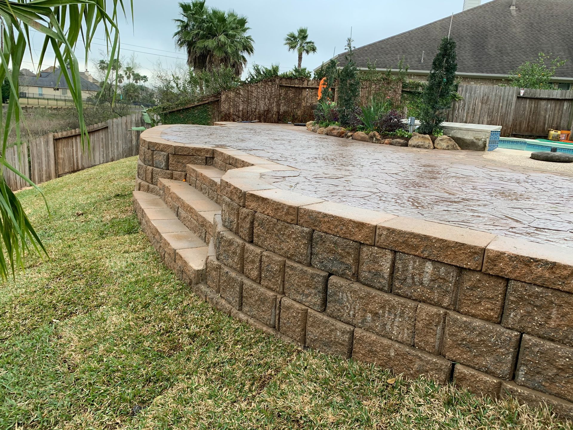Backyard patio with curved stone retaining wall, wet concrete surface, and palm trees by a pool