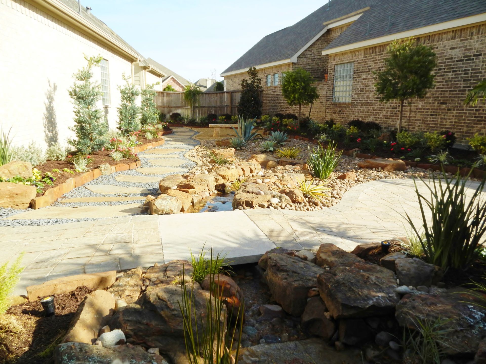 Stone-lined garden path and dry landscape between two houses under bright sunlight