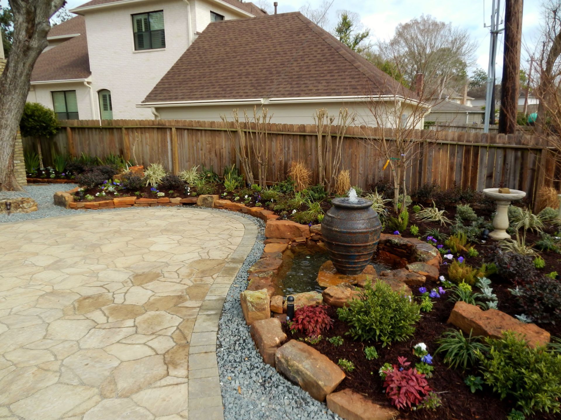 Curved stone patio beside a colorful landscaped garden and wooden fence in a backyard