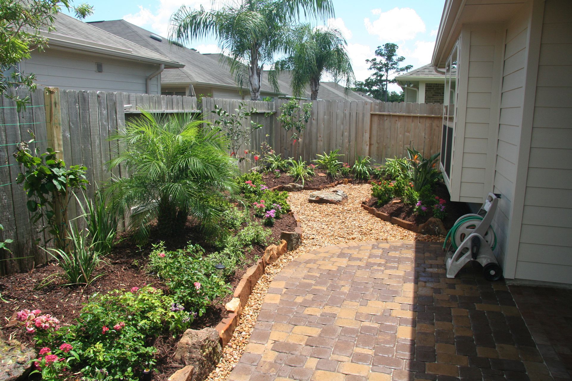 Backyard garden path with brick pavers, wooden fence, and flowering plants beside a house