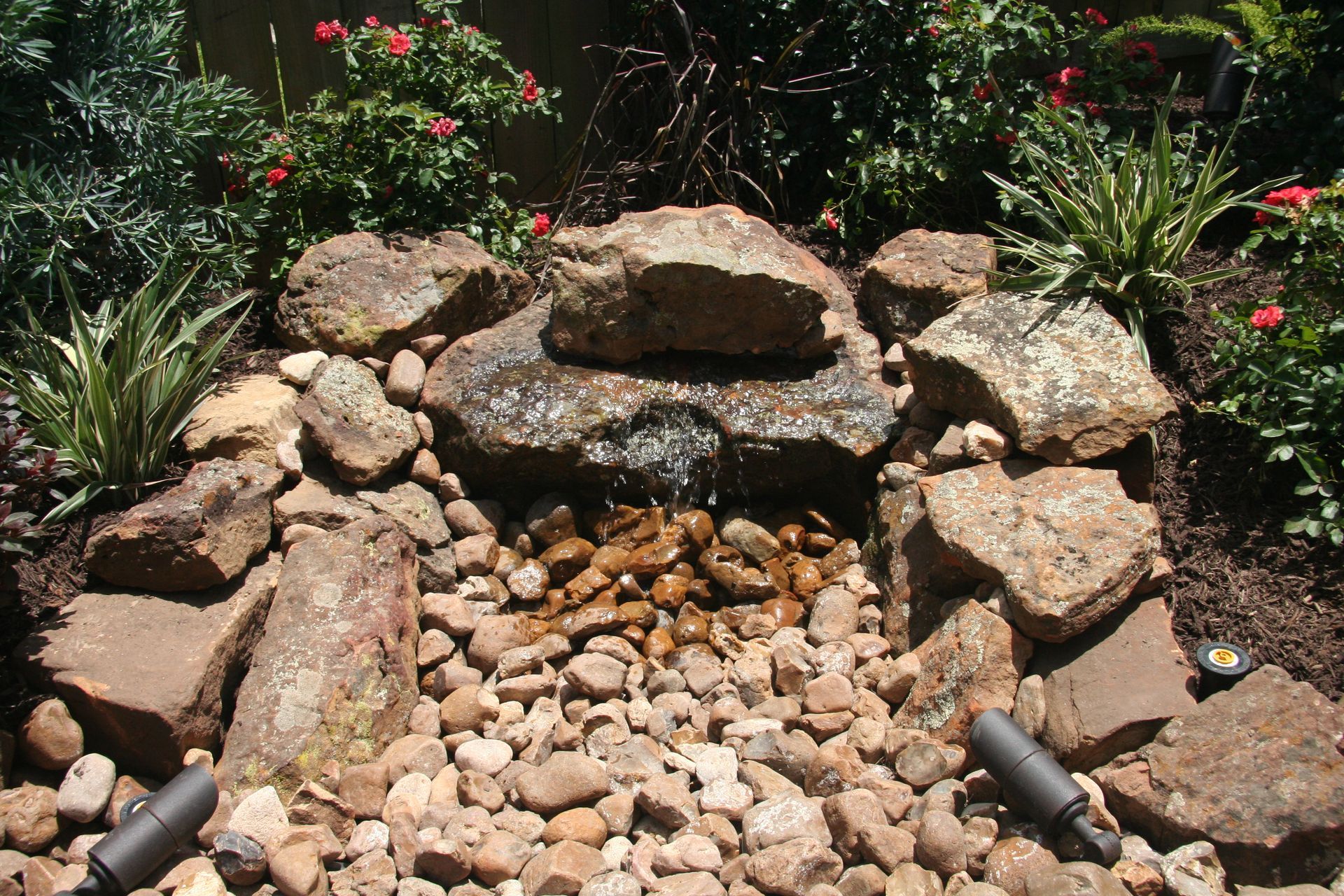 Rock garden with stacked stones and a dry creek bed of pebbles, surrounded by green plants and red flowers.