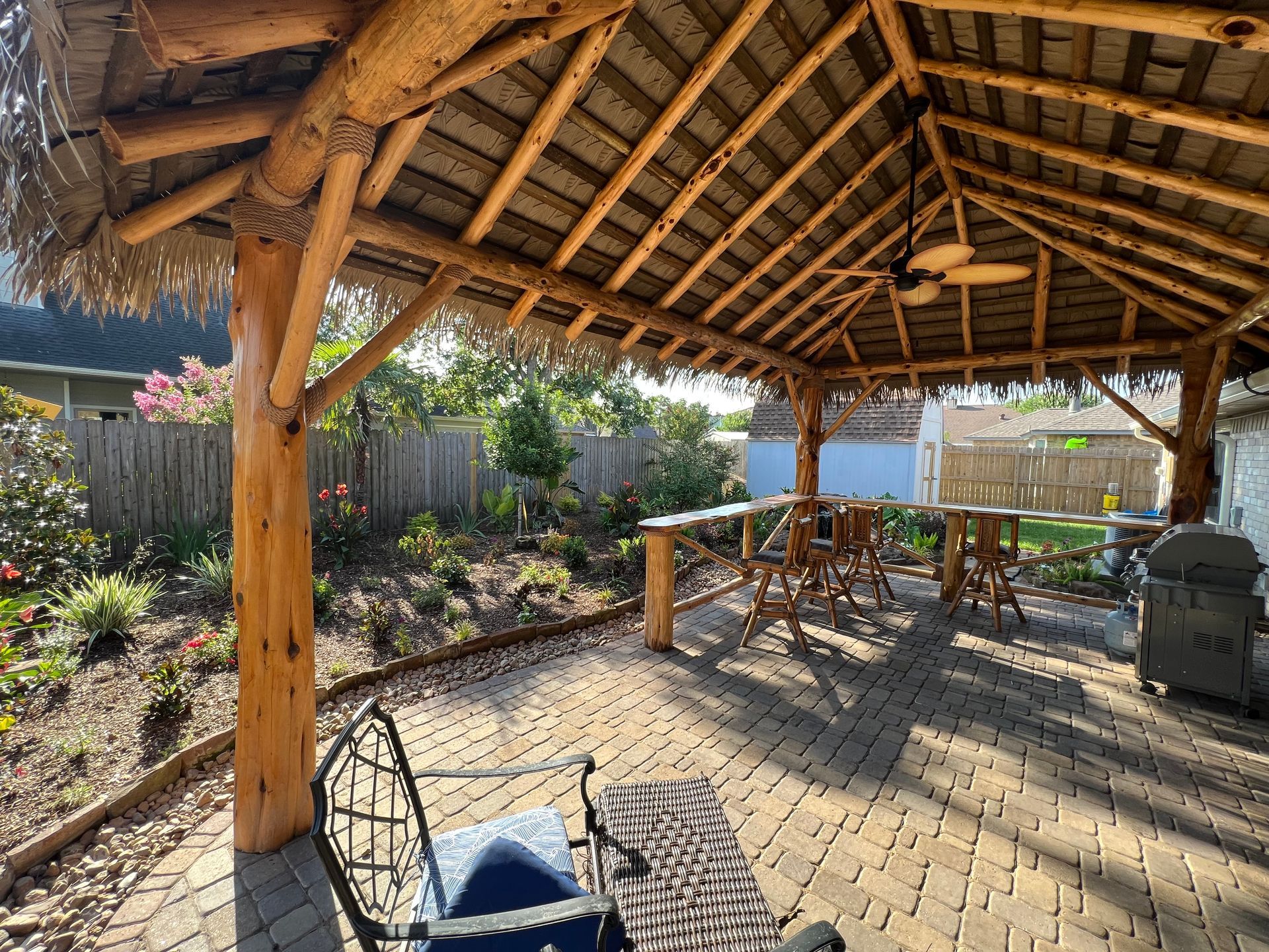 Covered outdoor patio with wooden beams, stone floor, table and chairs, and a grill in a sunny garden