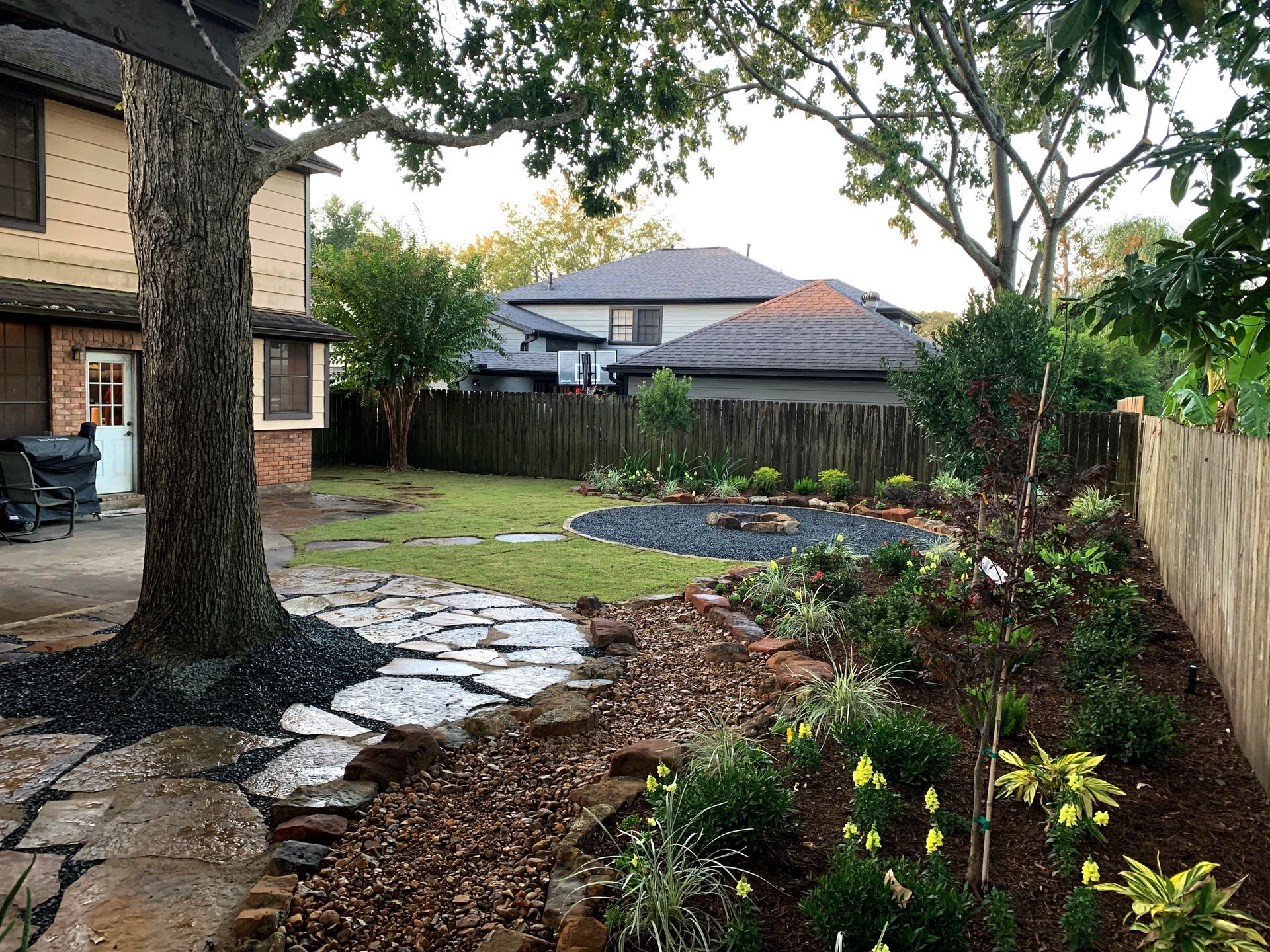 Backyard garden with stepping stones, mulched beds, grass, trees, and a wooden fence