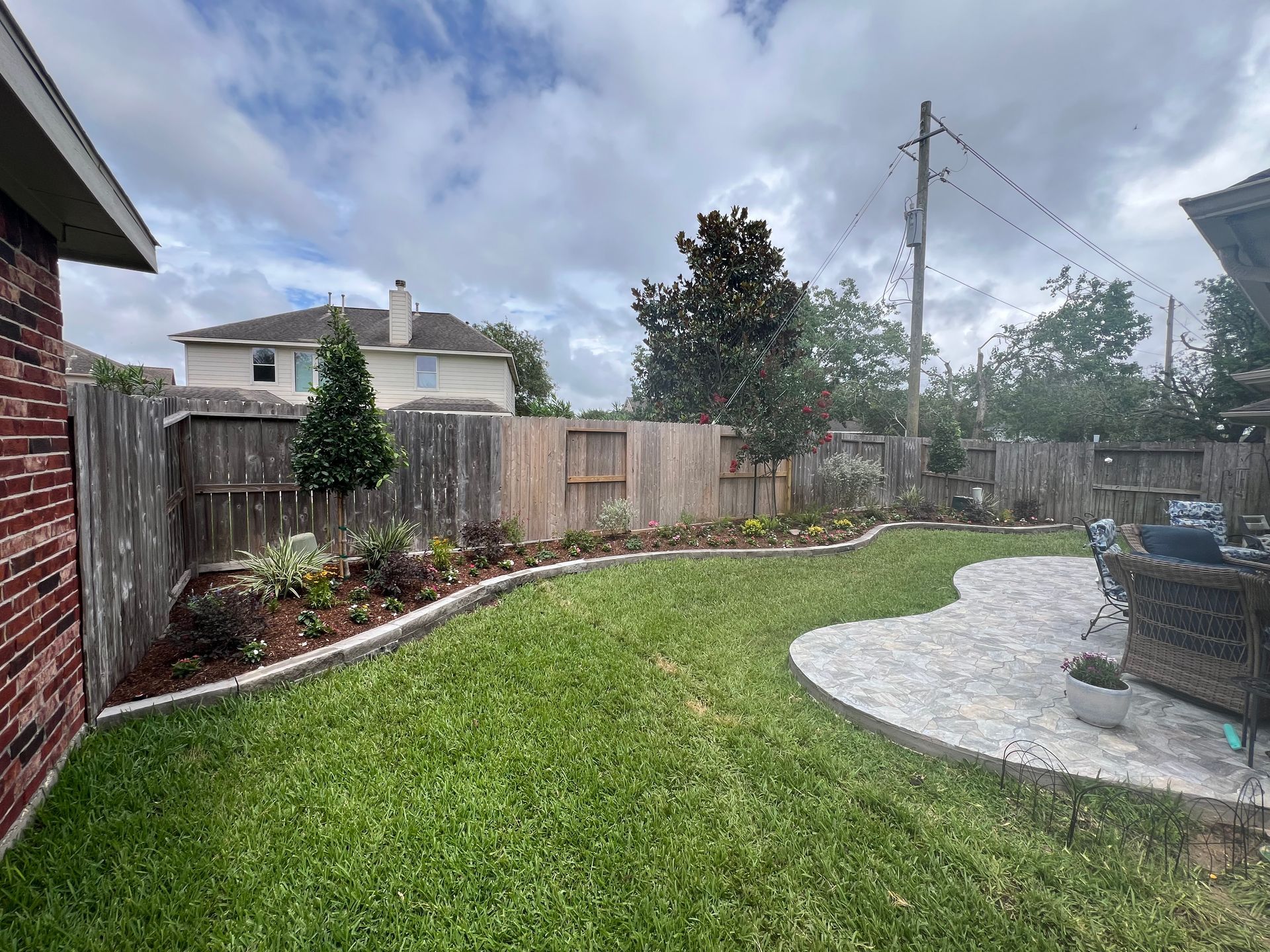 Backyard with green lawn, wooden fence, flower beds, and a curved patio with outdoor seating.