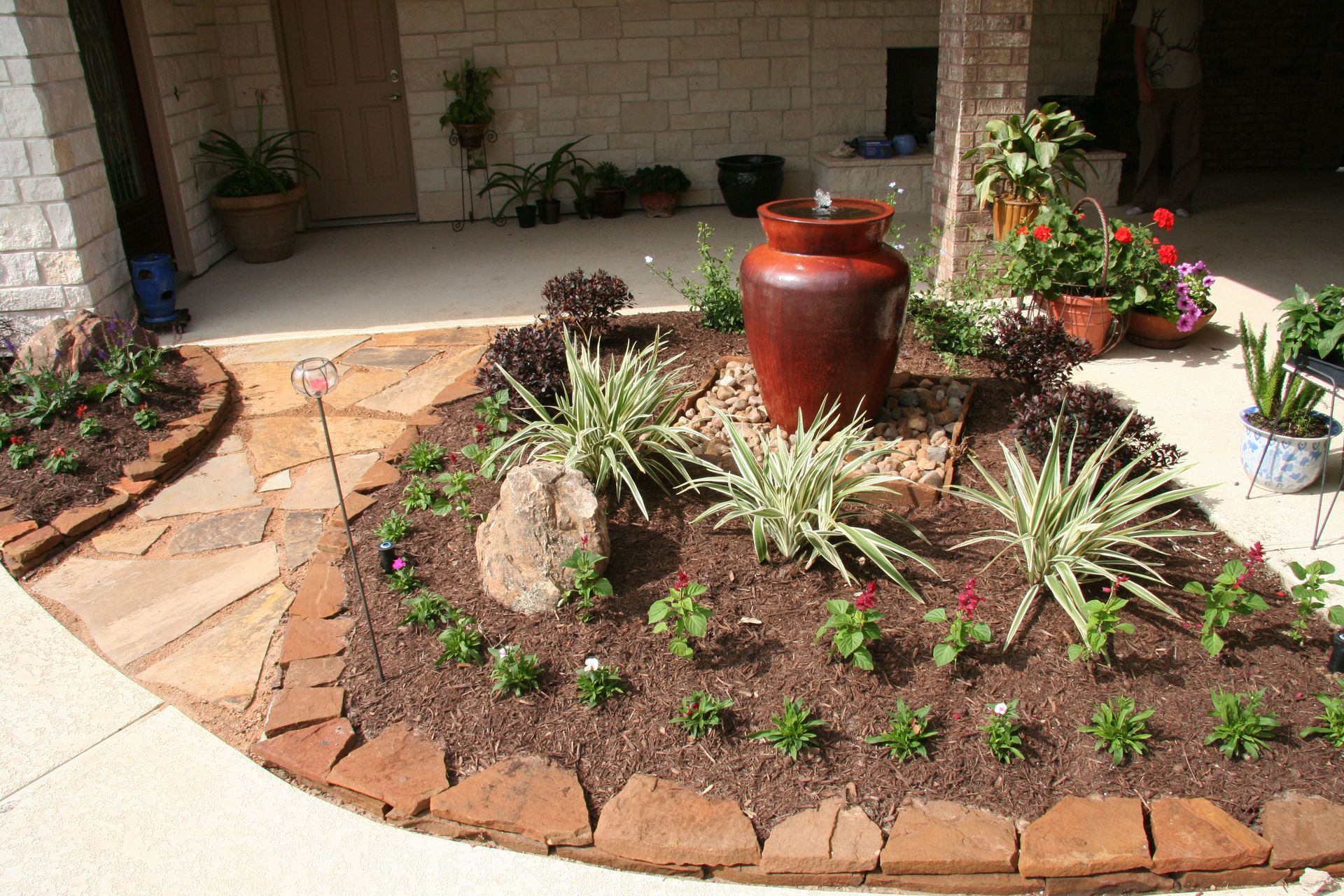 Front yard garden bed with red pottery, succulents, and brick edging against a house patio