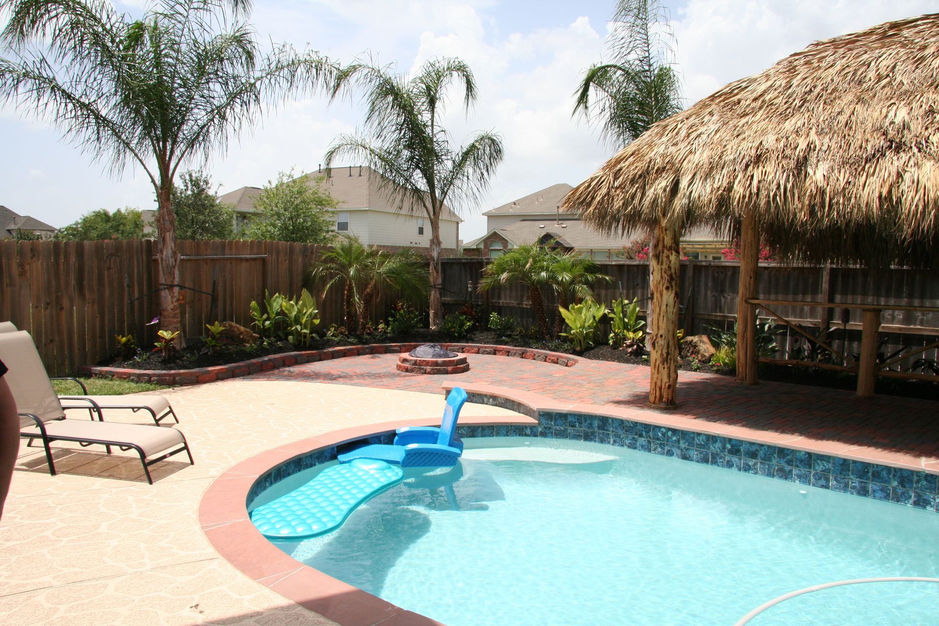 Backyard pool with palm trees, lounge chair, and thatched tiki bar in bright sunlight