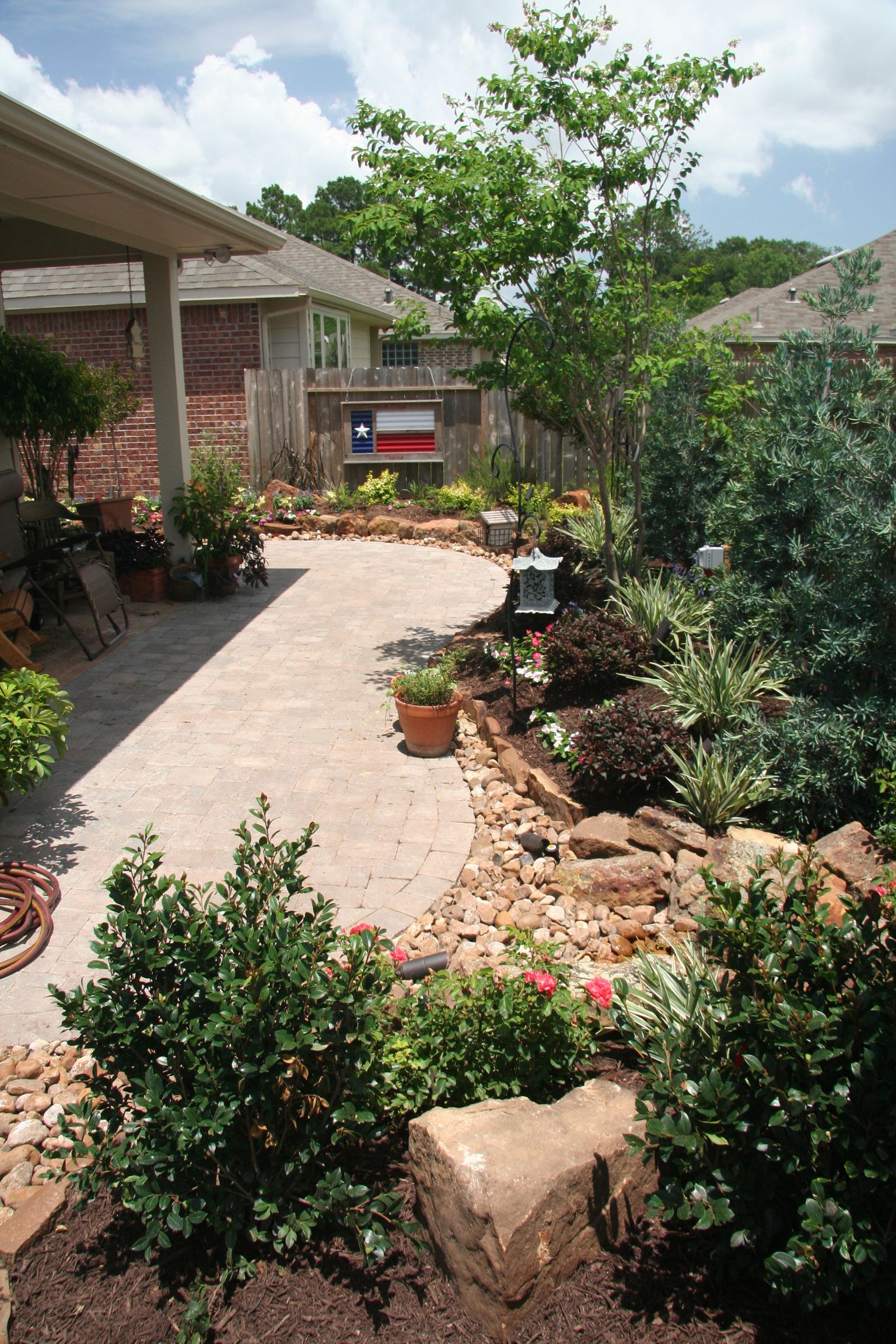 Sunny garden courtyard with potted plants, shrubs, and a stone pathway beside a house