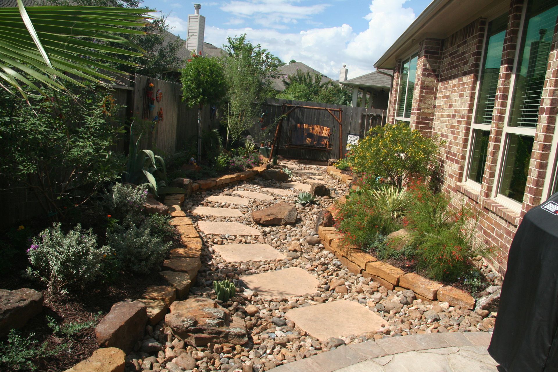 Narrow garden path with stepping stones and gravel beside a brick house and green plants